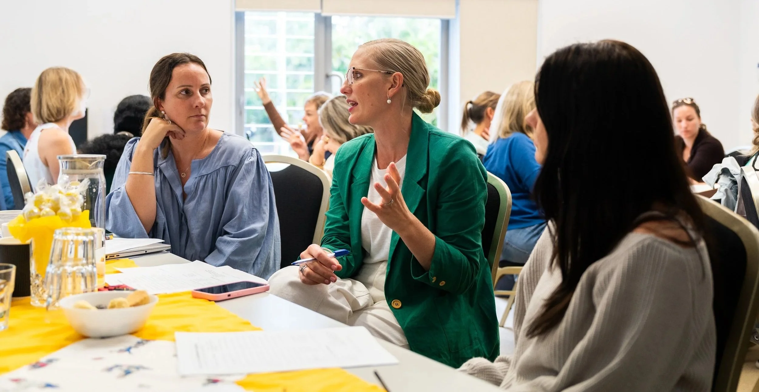 Women attending a discussion or meeting in a bright conference room with a window, some with papers and notebooks on the table, engaged in conversation.