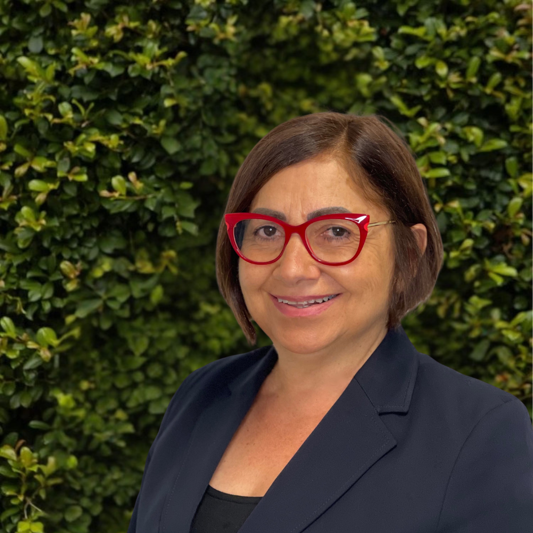 A woman with short brown hair, red glasses, wearing a black blazer, smiling, standing in front of green foliage.