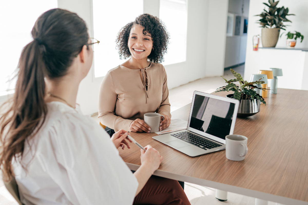 Two women engaging in a conversation at a wooden table with laptops and coffee mugs in a well-lit, modern office or home setting with plants.
