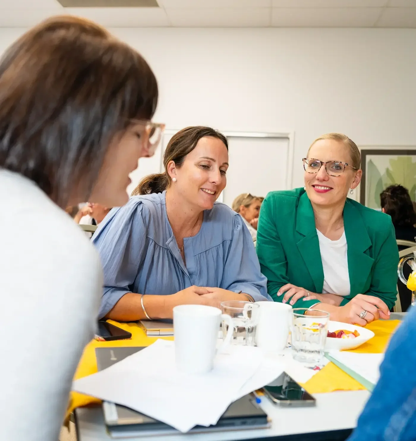 Three women sitting at a table engaged in conversation, with coffee mugs, glasses of water, notebooks, and a plate of food in front of them, in a well-lit room.