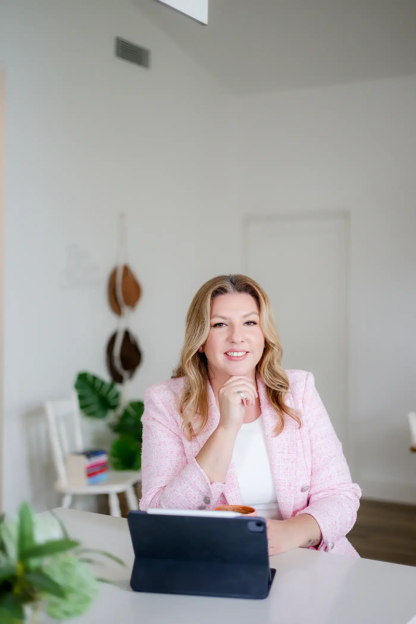 A woman with blonde hair wearing a pink blazer sitting at a table with her chin resting on her hand, smiling, in a bright room with plants, hanging hats, and a tablet on the table.