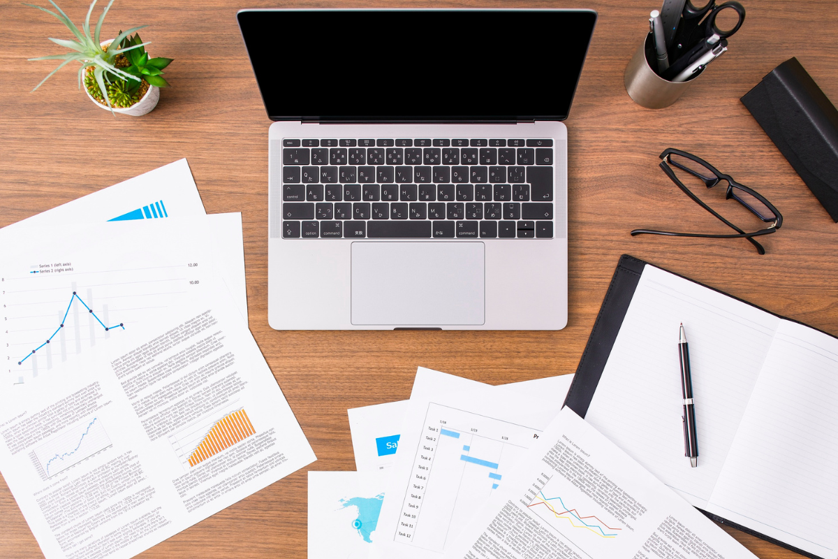 Work desk with a laptop, glasses, papers with graphs, a notebook, a pen, a pen holder, a black case, and a plant on a wooden surface.