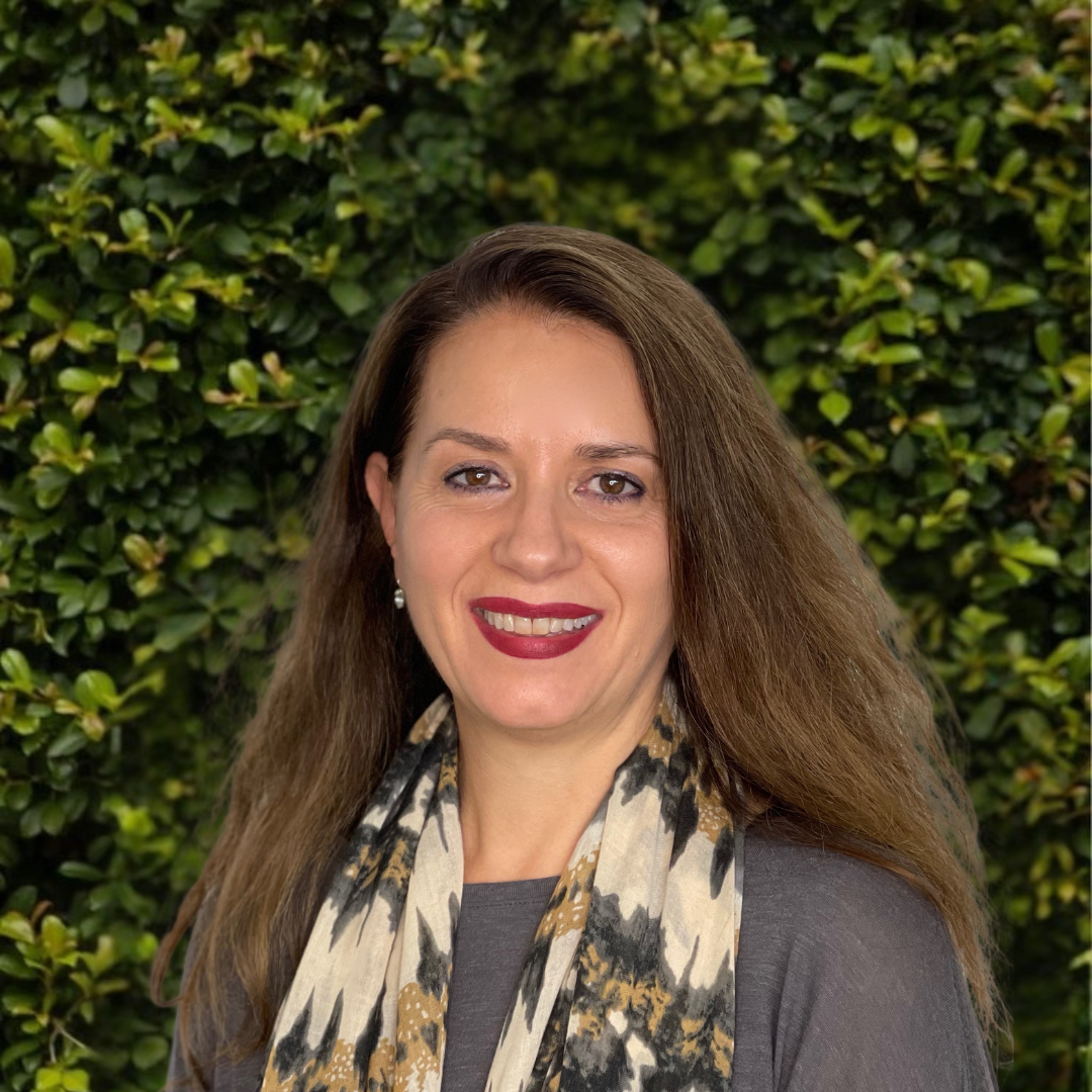A woman with long brown hair smiling in front of a green leafy background, wearing a gray top and patterned scarf.