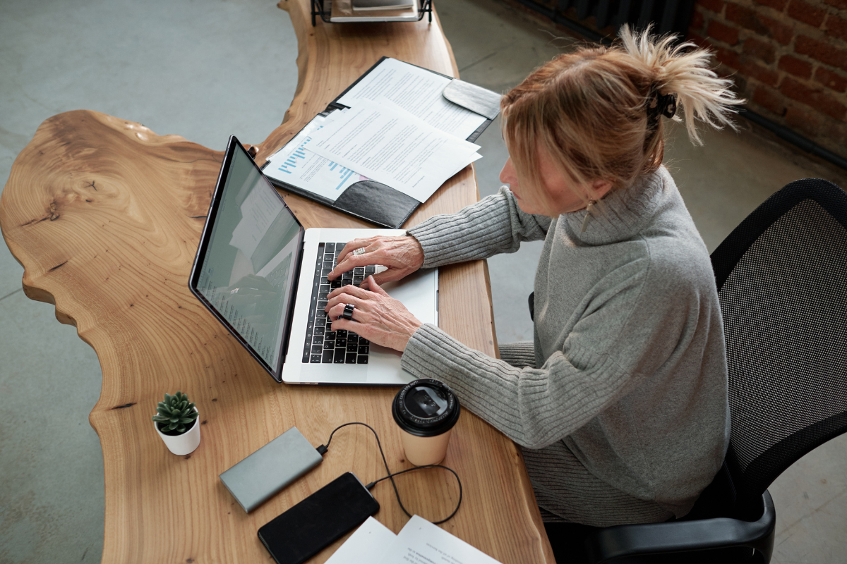 A woman with blonde hair tied in a ponytail sitting at a wooden desk working on a silver laptop. The desk has various papers, a small potted succulent, a black travel mug, and a portable charger. The background shows a brick wall and part of an office space.