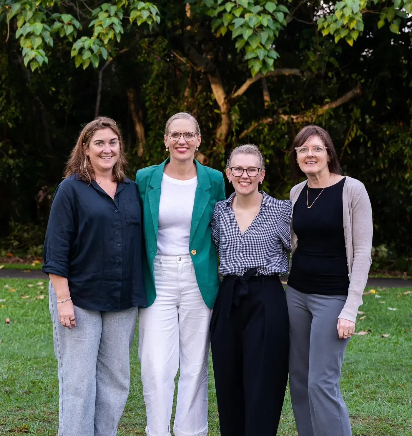 Four women standing outdoors on a grassy area with trees in the background, smiling at the camera.