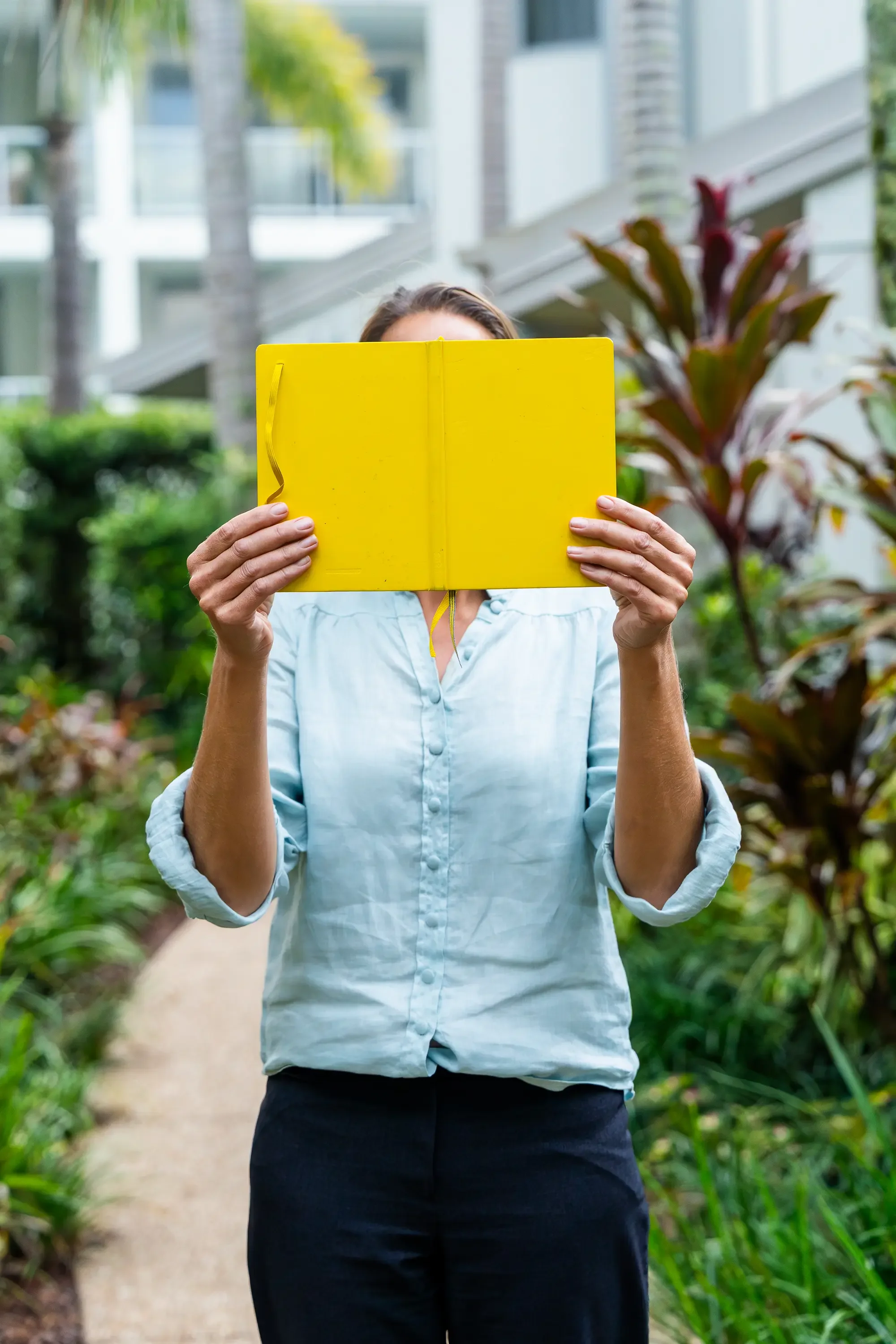 Person holding a yellow notebook in front of face, standing on a garden path with green plants and trees in background.
