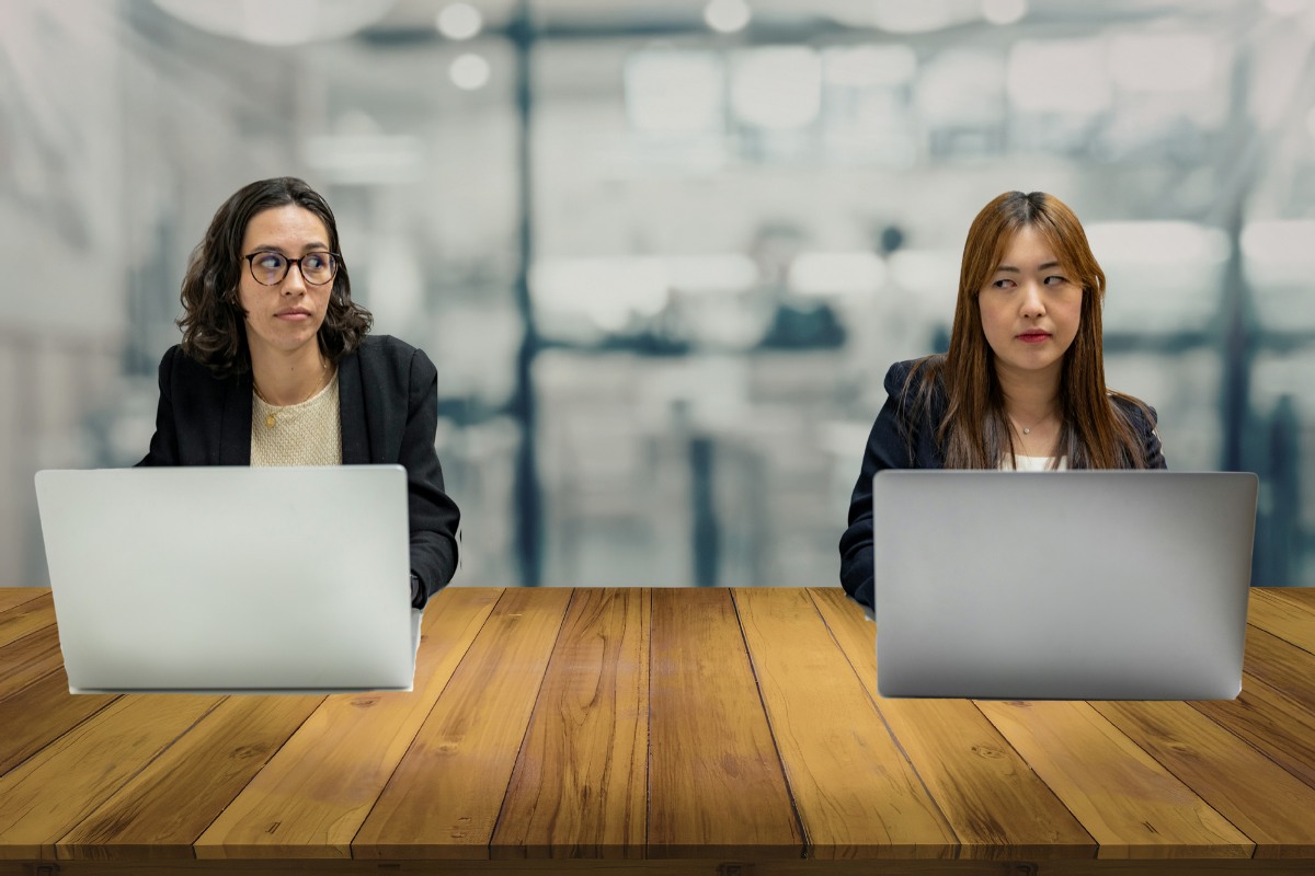 Two women sitting at a wooden table with laptops in front of them in a modern office environment, one on the left with glasses and the other on the right with long brown hair.