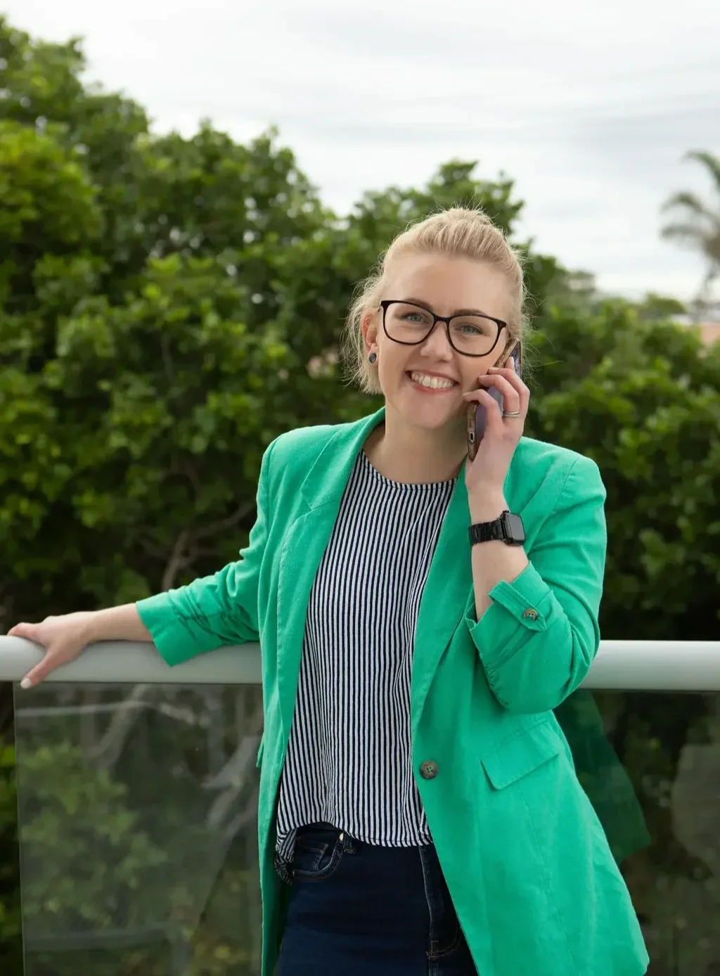 A woman with glasses and blonde hair, smiling while talking on her cellphone outdoors, wearing a turquoise blazer and striped shirt.