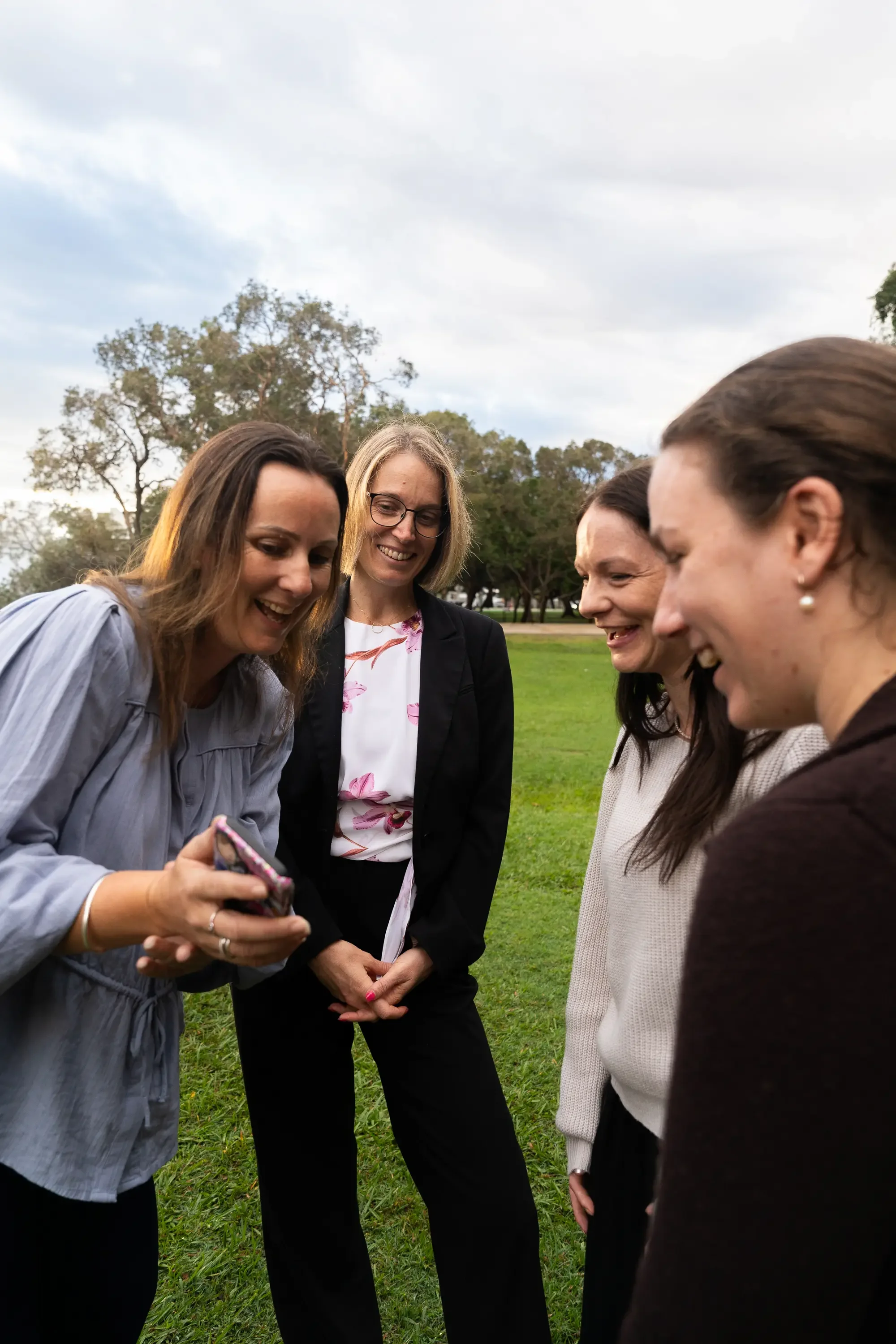 Four women standing outdoors on a grassy field, looking at a smartphone held by one woman, smiling and engaging with each other, with trees and a cloudy sky in the background.
