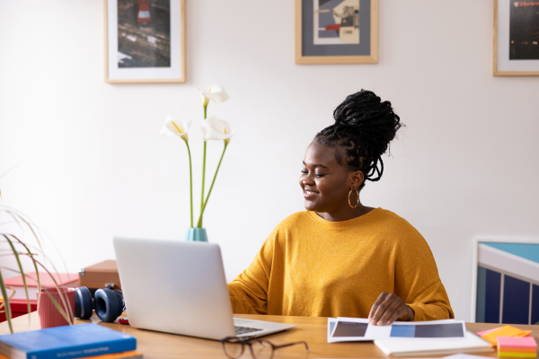 woman in yellow top smiling at her laptop screen, headphone, books, glasses on desk, art on walls behind. Casual work-from-home setting.