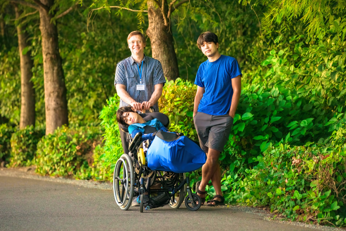 Two men standing outdoors next to a child in a wheelchair, with lush greenery and trees in the background.