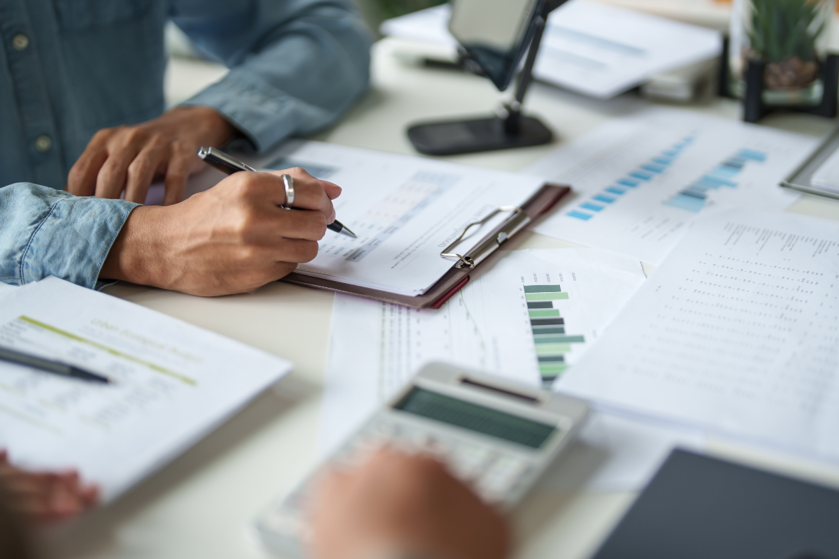 People working on financial or business documents with graphs, charts, a calculator, and a computer on a desk.