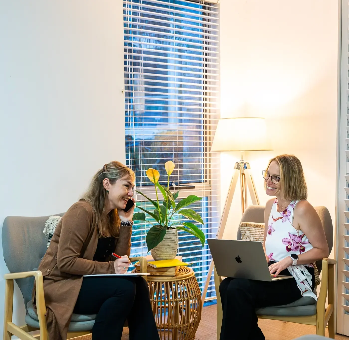 Two women sitting in a brightly lit room, engaging in a lively conversation. One woman, with long brown hair, is on the phone, smiling, with a notebook and pen in her hands. The other woman, with shoulder-length blonde hair and glasses, is smiling at her laptop, which is on her lap. There is a large window with blinds behind them, a potted plant on a small table between them, and a standing lamp providing cozy lighting.