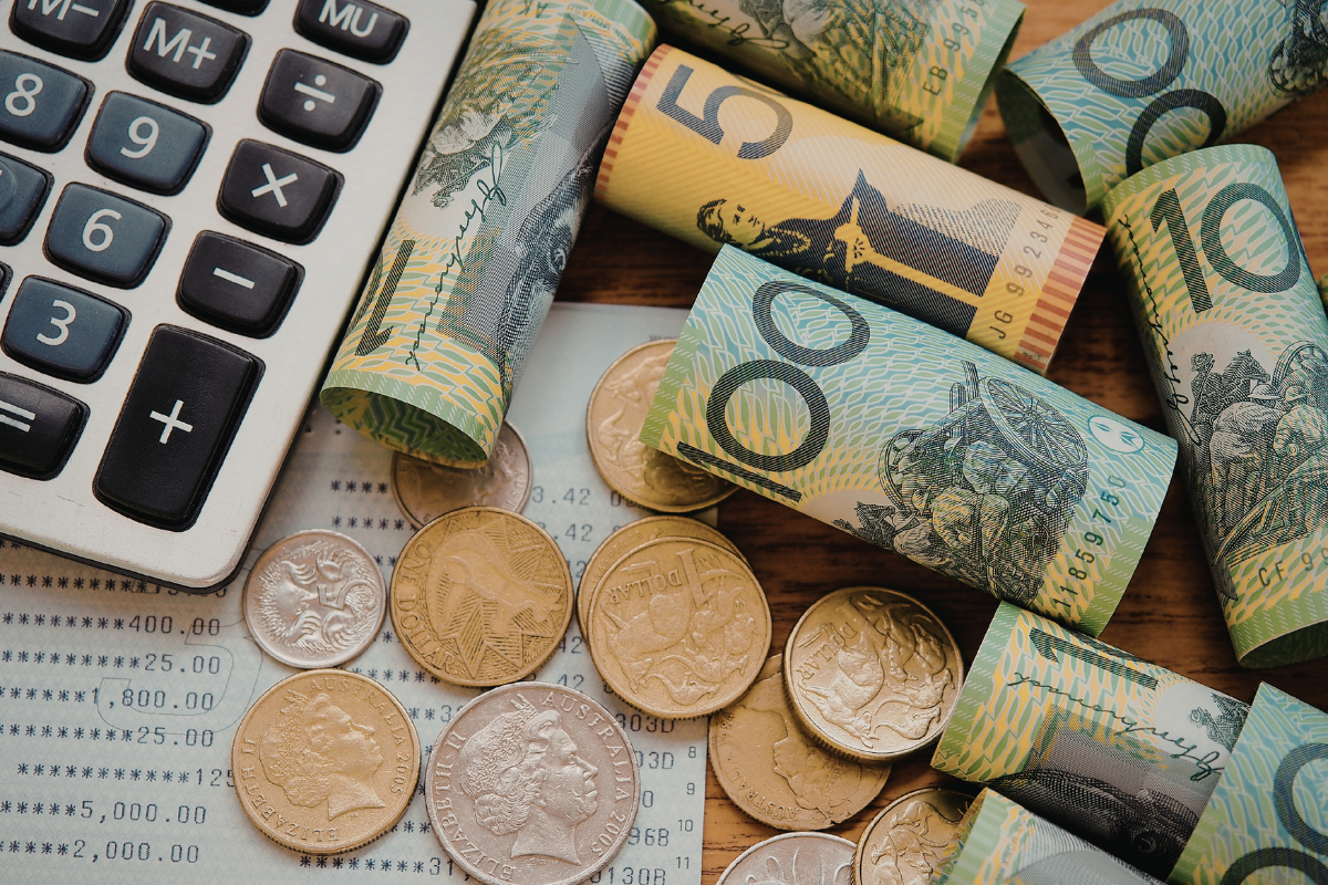 Australian banknotes in denominations of 5, 10, and 50 dollars, rolled up and scattered on a wooden surface, with coins and a calculator and a bank statement.