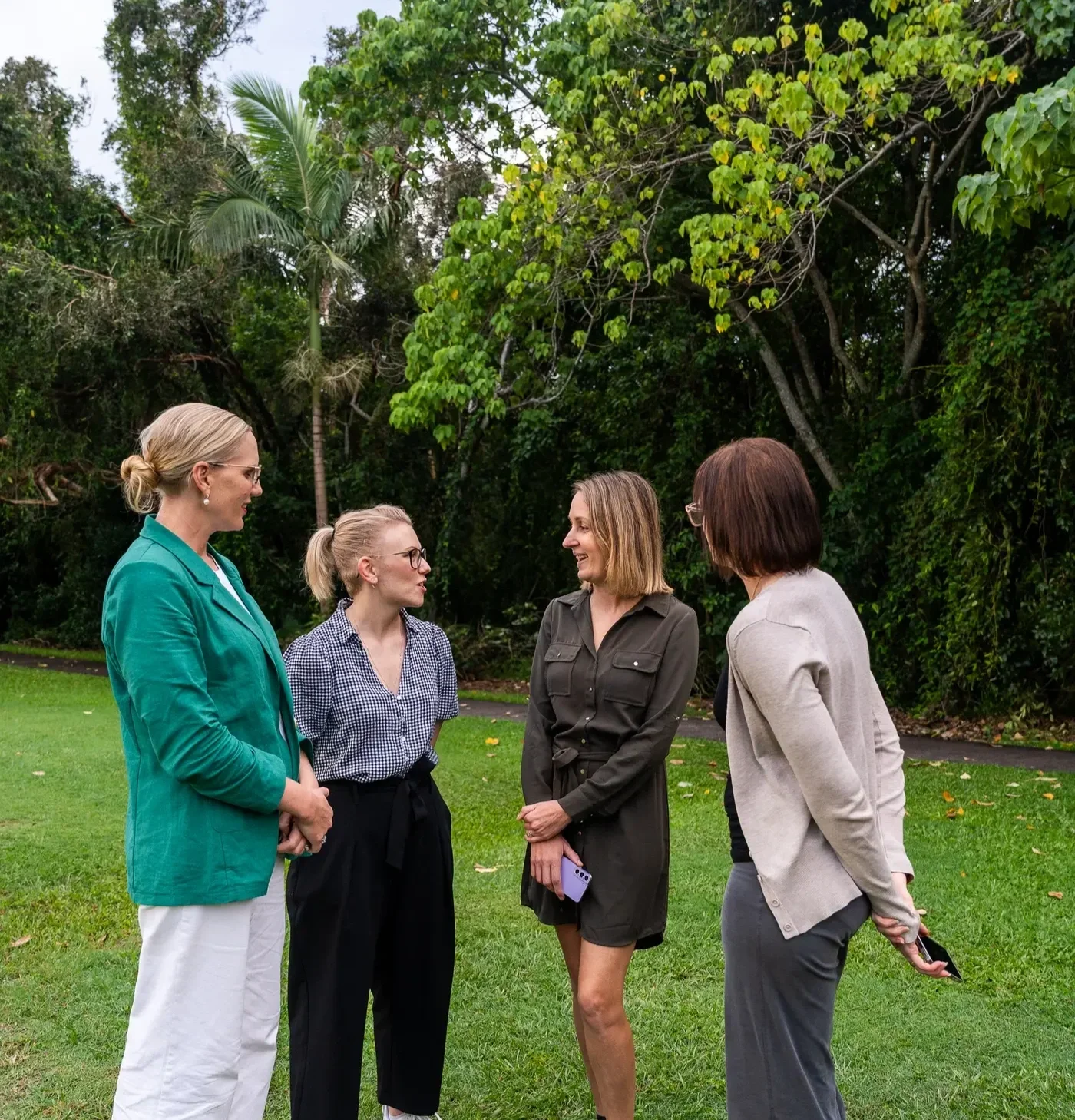 Four women standing and talking outdoors on grass, with trees in the background.