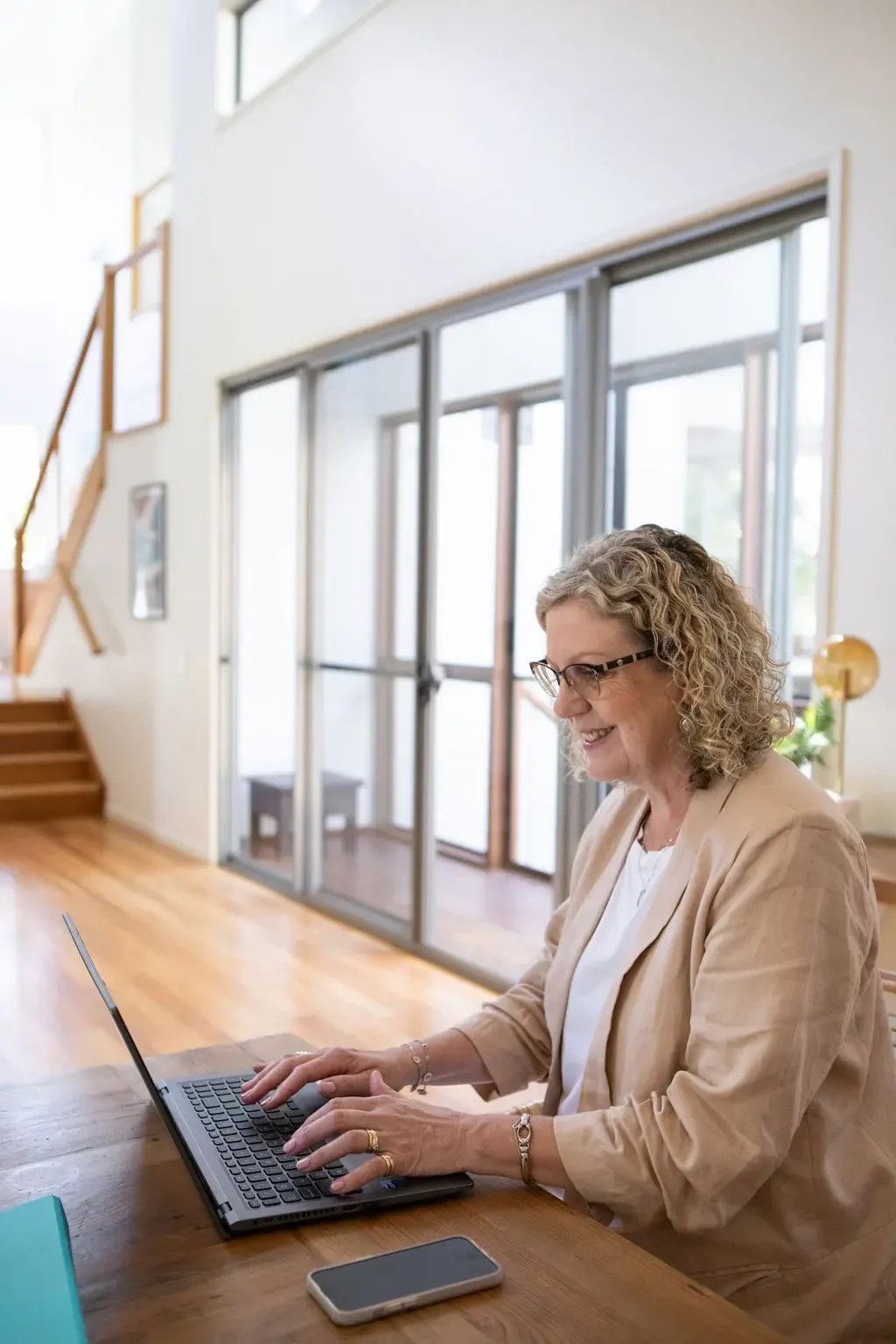 A woman with glasses sitting at a wooden table working on a laptop in a bright, modern home interior with large windows and wooden stairs.