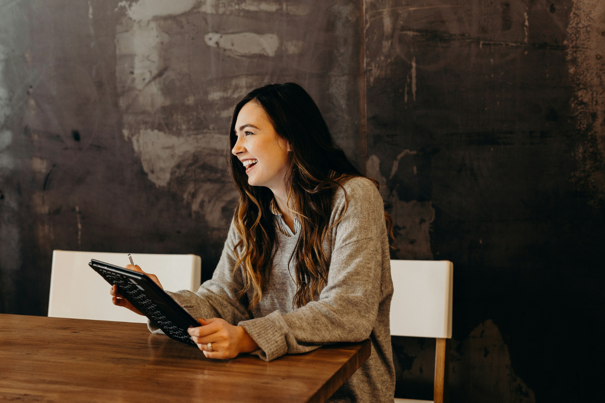 A woman with long wavy hair smiling and holding a tablet, sitting at a wooden table in front of a dark textured wall.