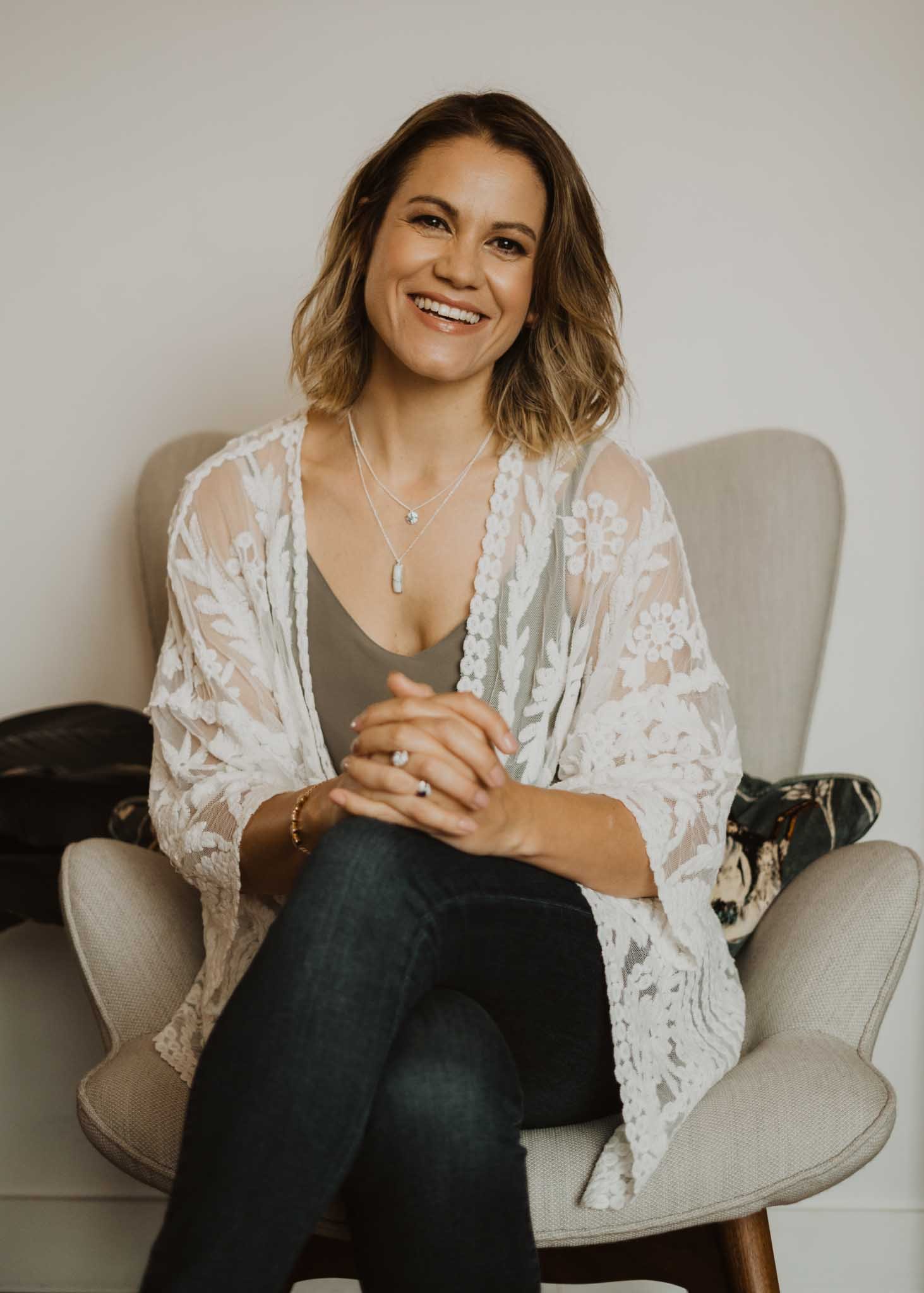 A woman with shoulder-length wavy brown hair, smiling, sitting in a beige armchair, wearing a gray top, a white lace cardigan, black pants, and layered necklaces.