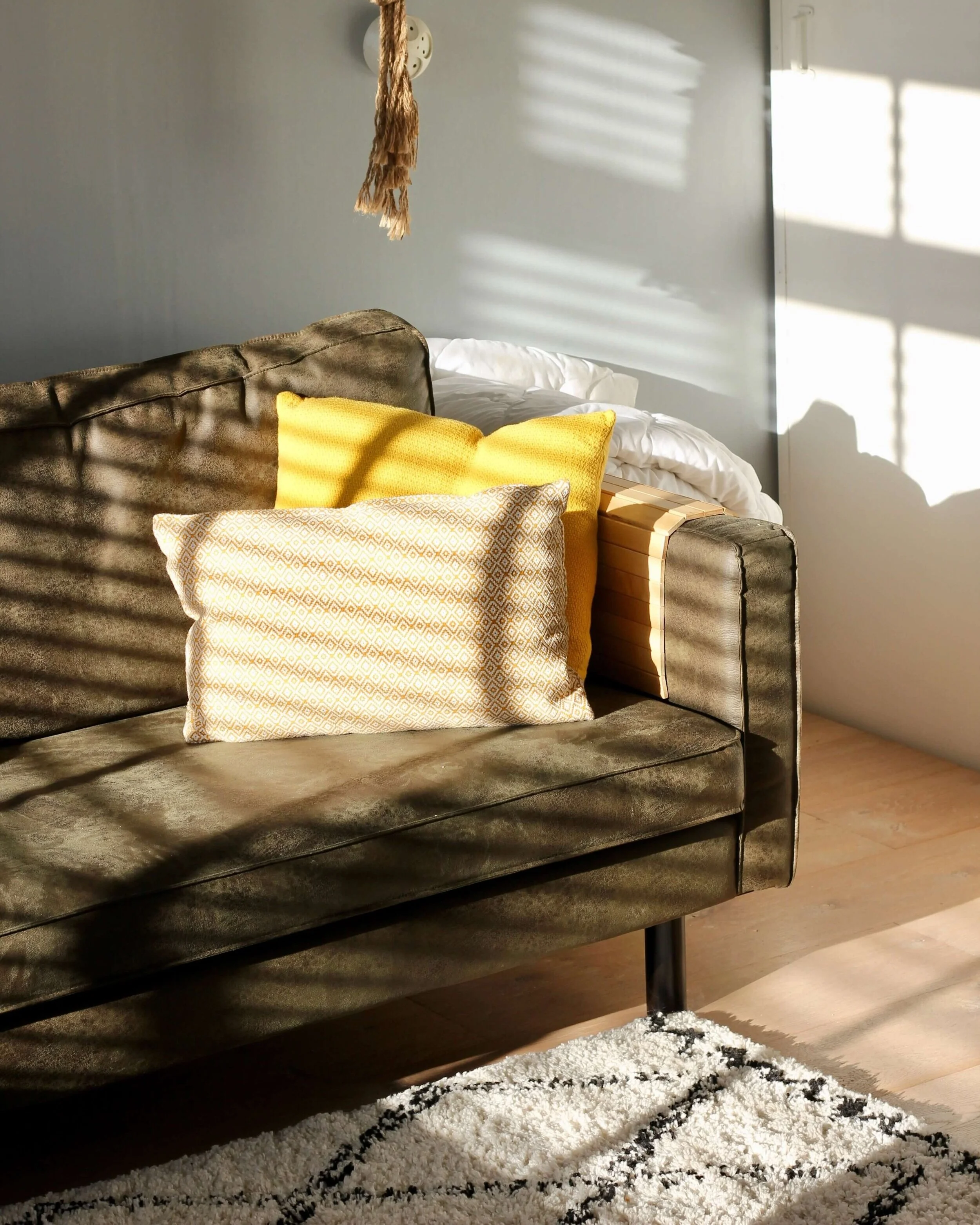 A gray sofa with yellow pillows, a wooden side table, and a black and white patterned rug in a room with sunlight and shadows on the wall.