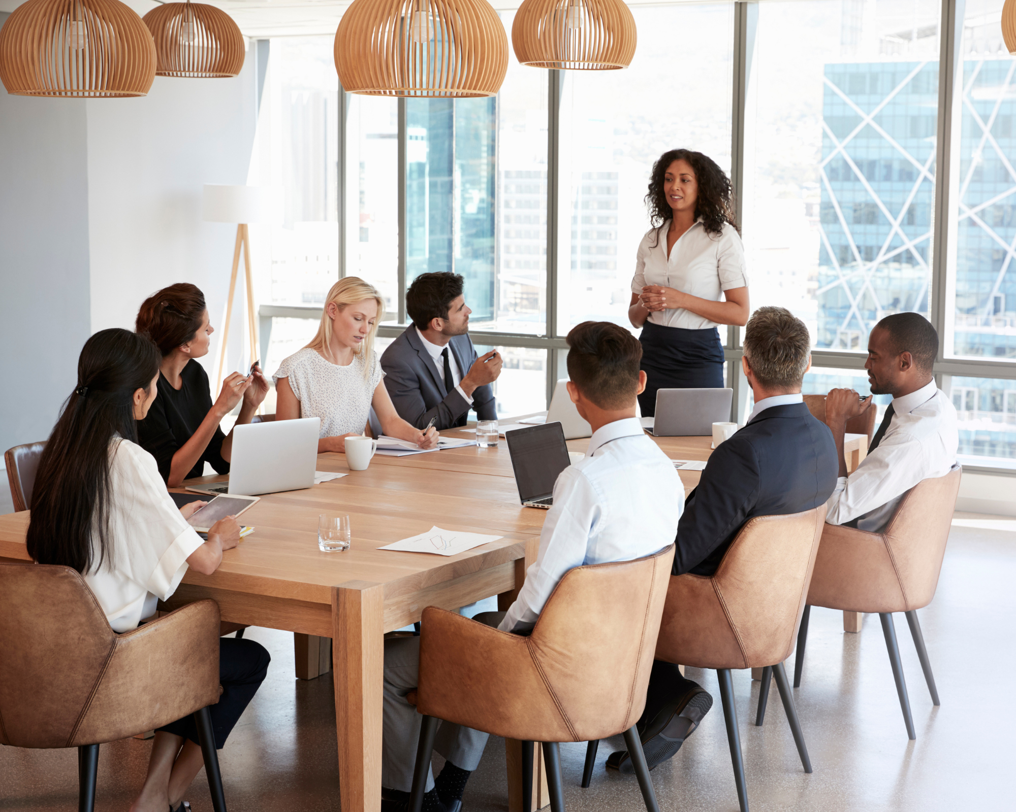 A diverse group of professionals in a modern office meeting room, sitting around a large wooden table with laptops and notebooks. A woman with curly hair is standing and speaking to the group, with large windows showing city buildings in the background.