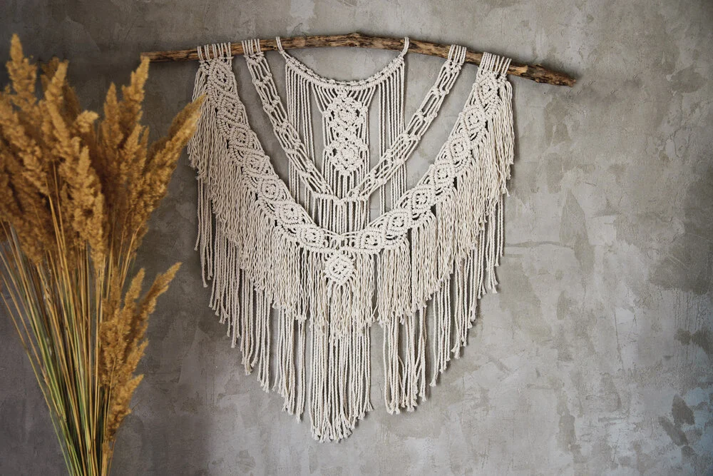 Macrame wall hanging on a wooden stick, with fringe details, hanging on a gray wall, next to dried tan pampas grass.