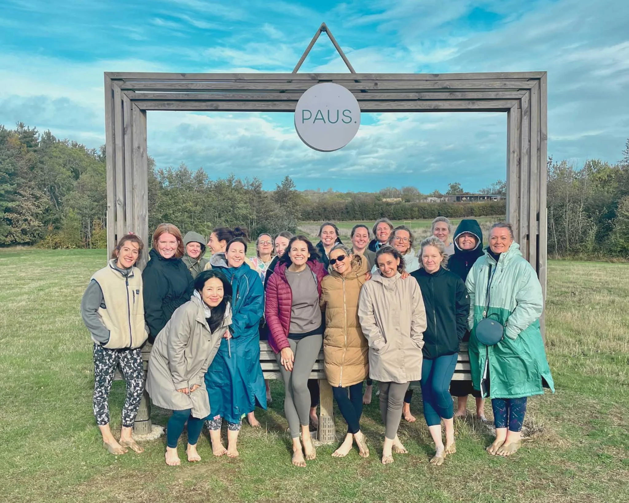 Group of people standing outdoors on grass, smiling, wearing jackets and casual clothing, beneath a wooden frame with a round sign reading 'PAUS' hanging above them, with trees and a partly cloudy sky in the background.