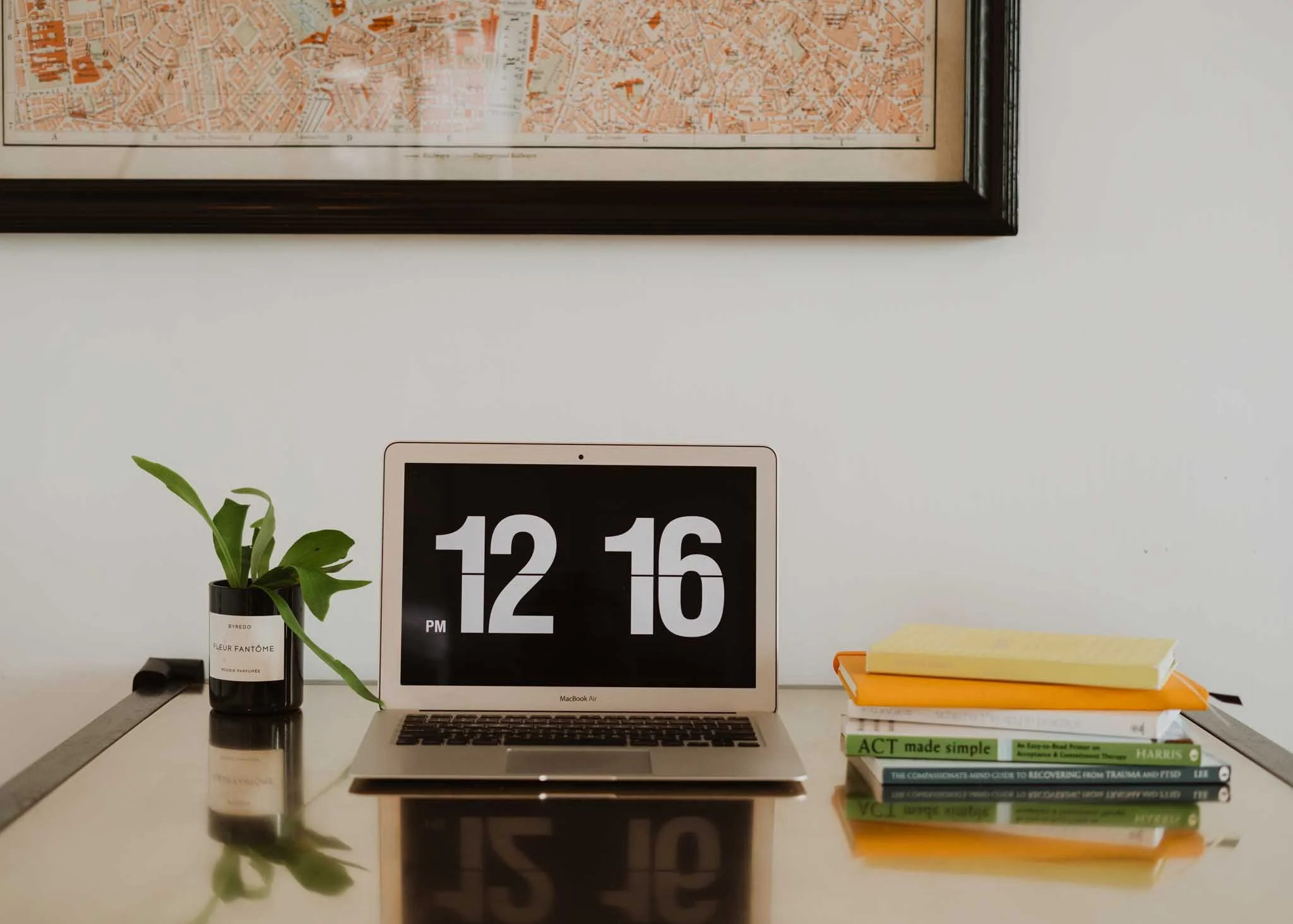 A workspace with a silver MacBook Air laptop displaying the time 12:16 PM, a potted green plant, a white clock, and a stack of yellow and green books on a white desk, with a framed map on the wall behind.
