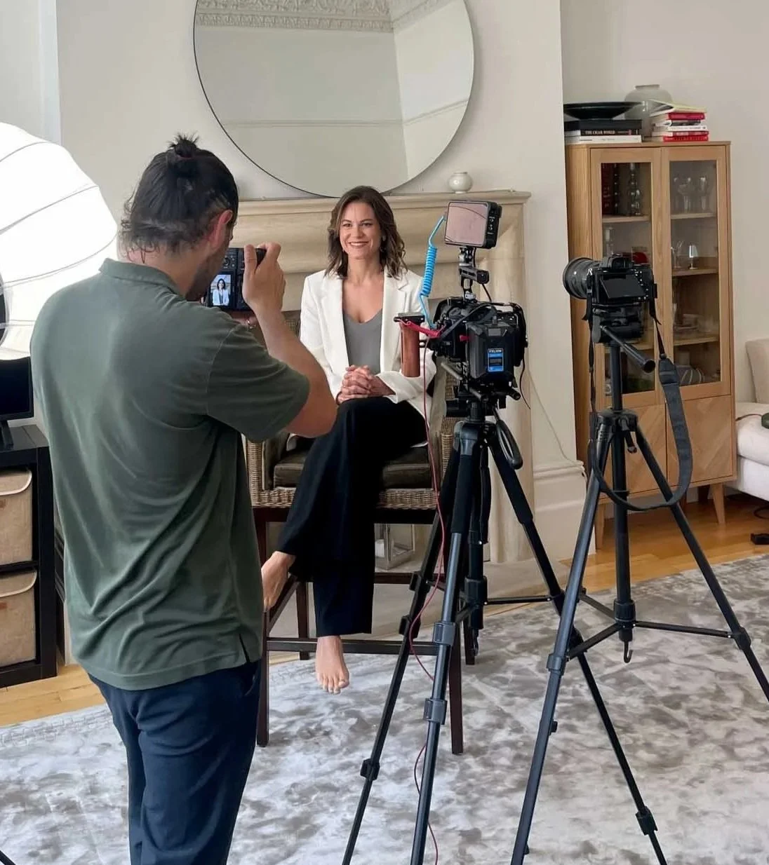 A woman sits on a chair in front of a camera and lights, smiling for a photo or video shoot, with a man taking her picture or recording her, in a cozy room with wooden furniture and a large mirror.