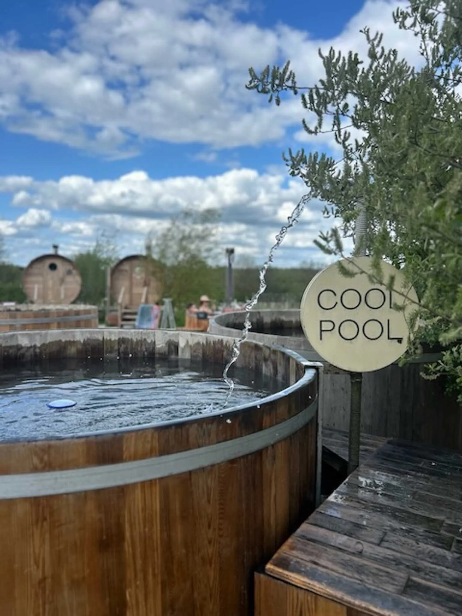 A hot tub with water bubbling inside, surrounded by a wooden deck, with a sign that reads 'COOL POOL'. In the background, there are holiday trailers, trees, and a partly cloudy sky.