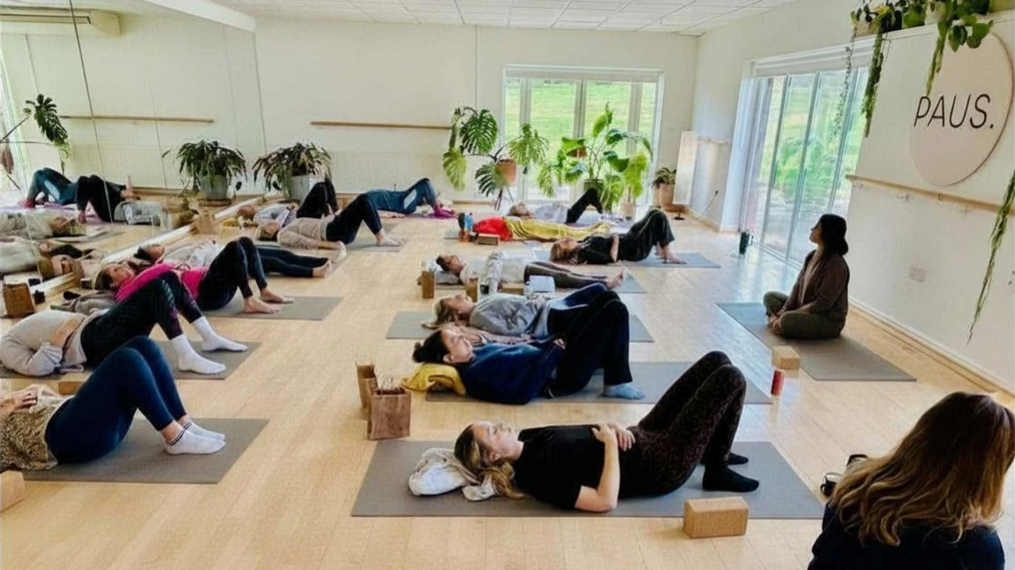 People participating in a yoga class, lying on yoga mats in a bright studio with large windows, plants, and a wooden floor, with an instructor sitting cross-legged at the front.