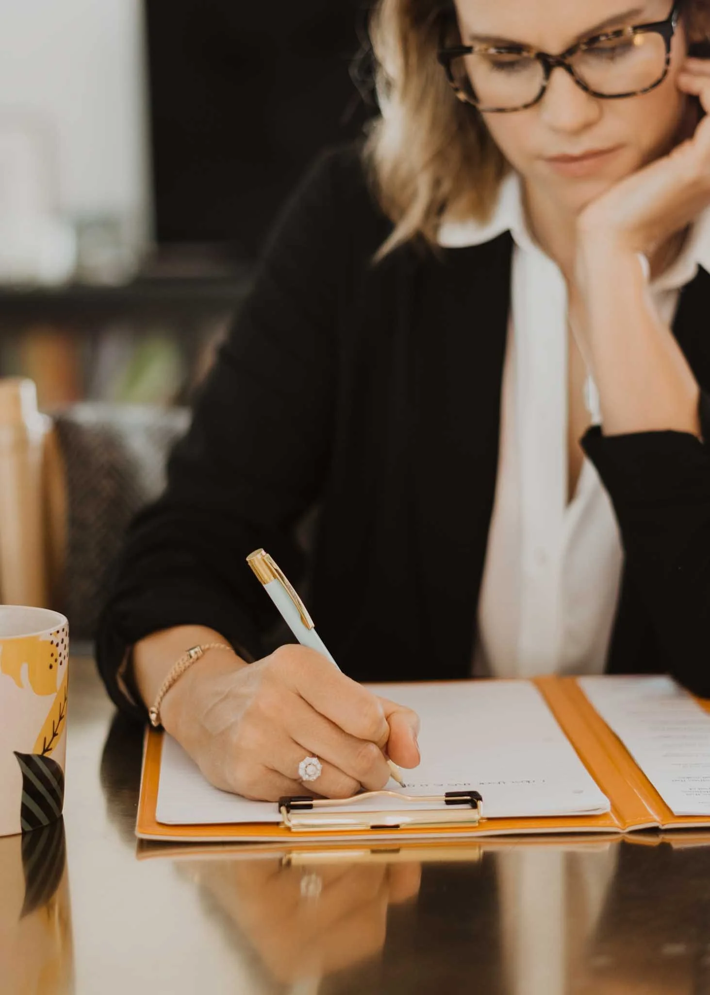 A woman with glasses and blonde hair, wearing a white shirt and black sweater, is sitting at a desk writing in a notebook with a gold and white pen. There is a mug with yellow design on the side in the foreground, and she is resting her head on her left hand.