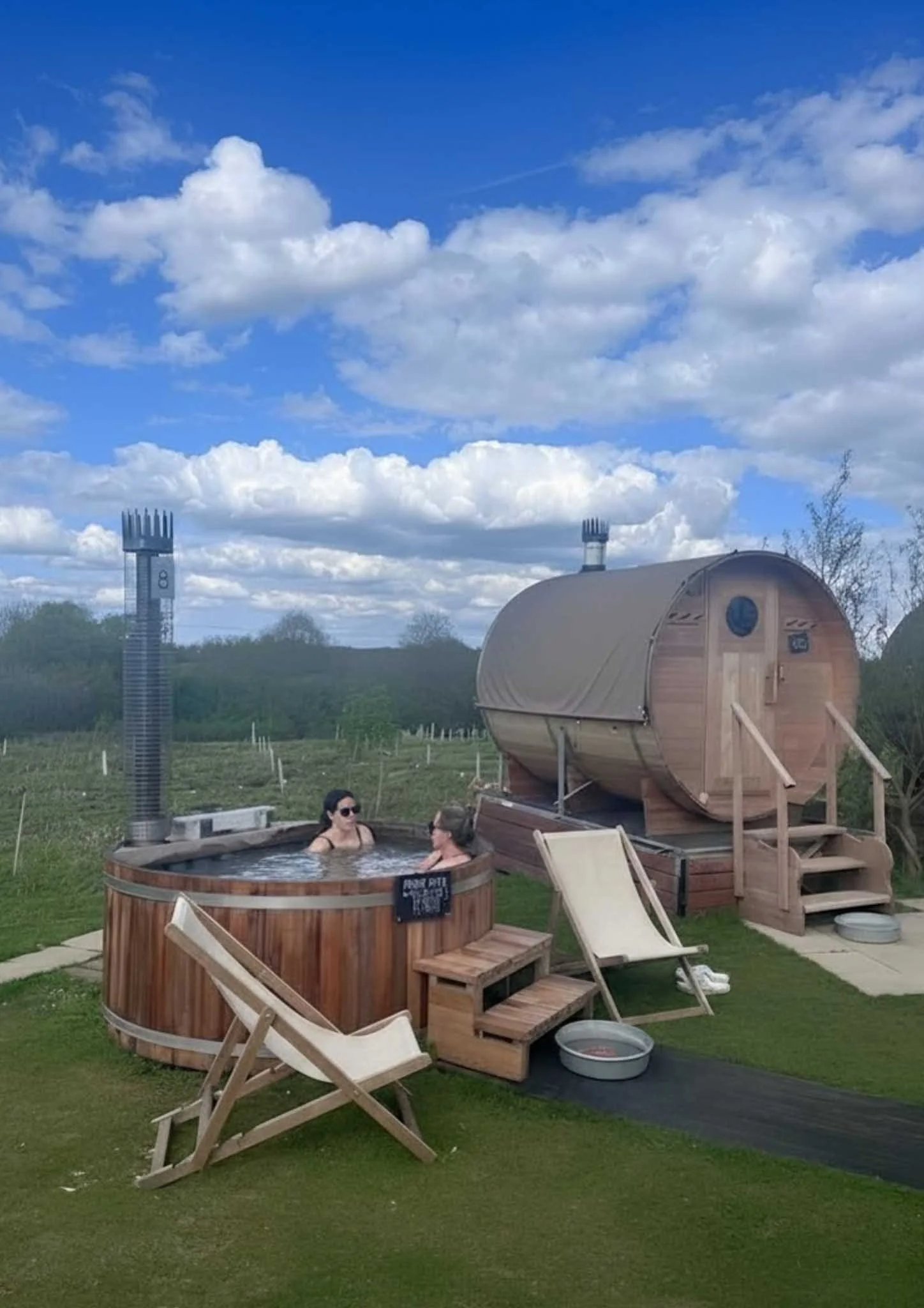 Two women relaxing in a round hot tub outdoors near a wooden sauna with stairs, chairs, and a black walkway, under a blue sky with scattered clouds.