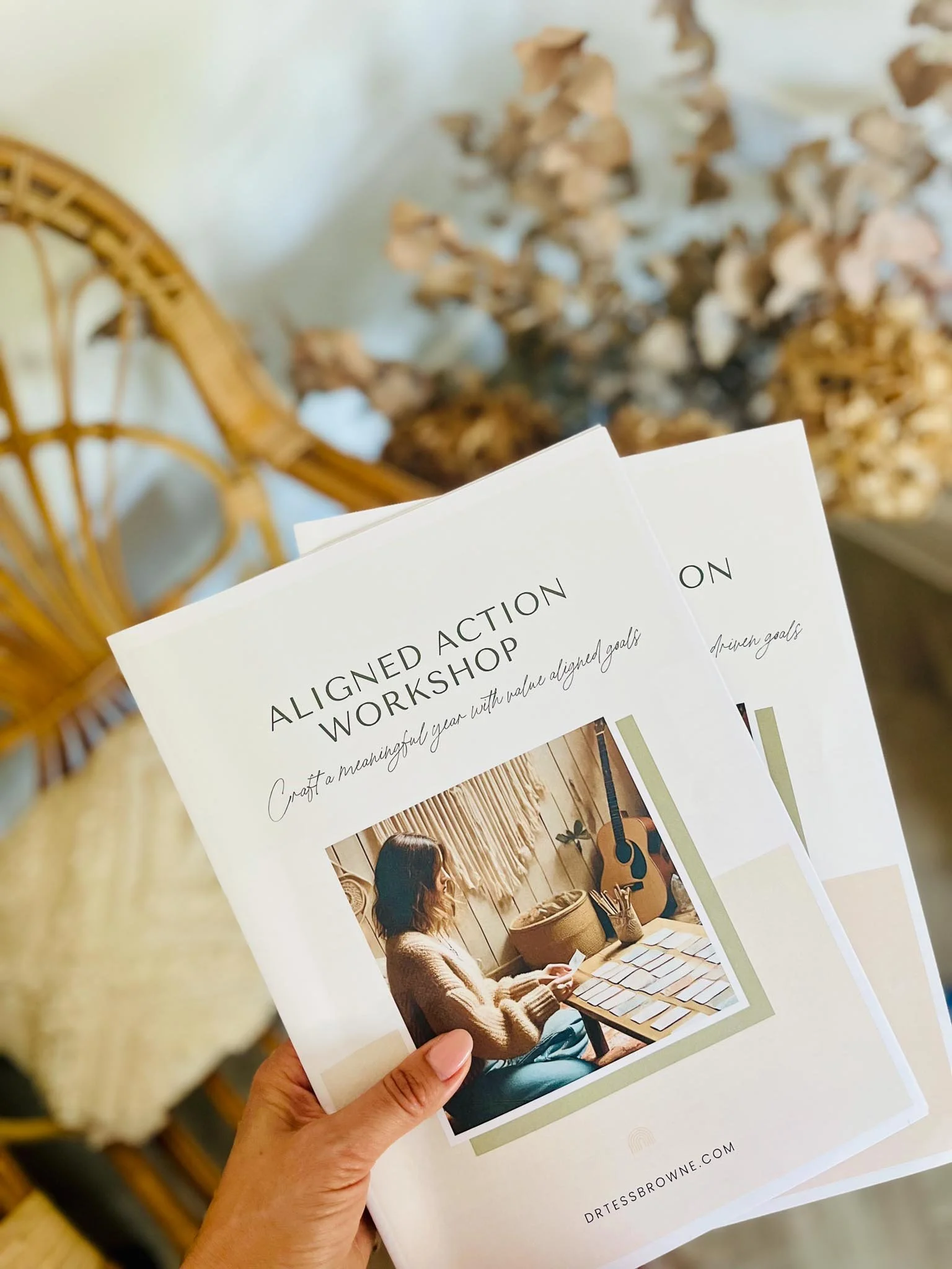 A hand holding a flyer for an 'Aligned Action Workshop' with a photo of a woman sitting at a table with color swatches and a guitar in a cozy room.