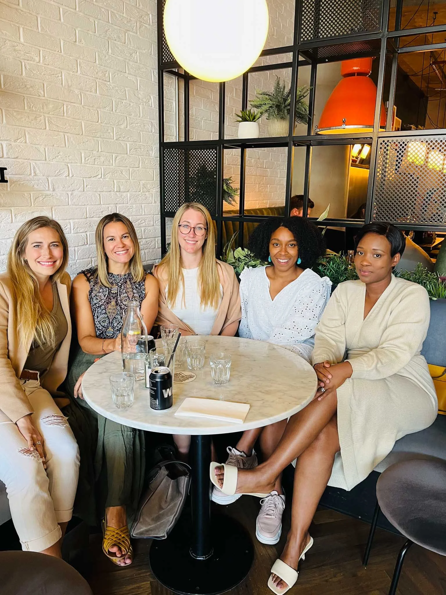 Group of five women sitting at a round table in a restaurant, smiling at the camera. The table has glasses, a can, and a water bottle. The background features a white brick wall, plants, and decorative lighting.