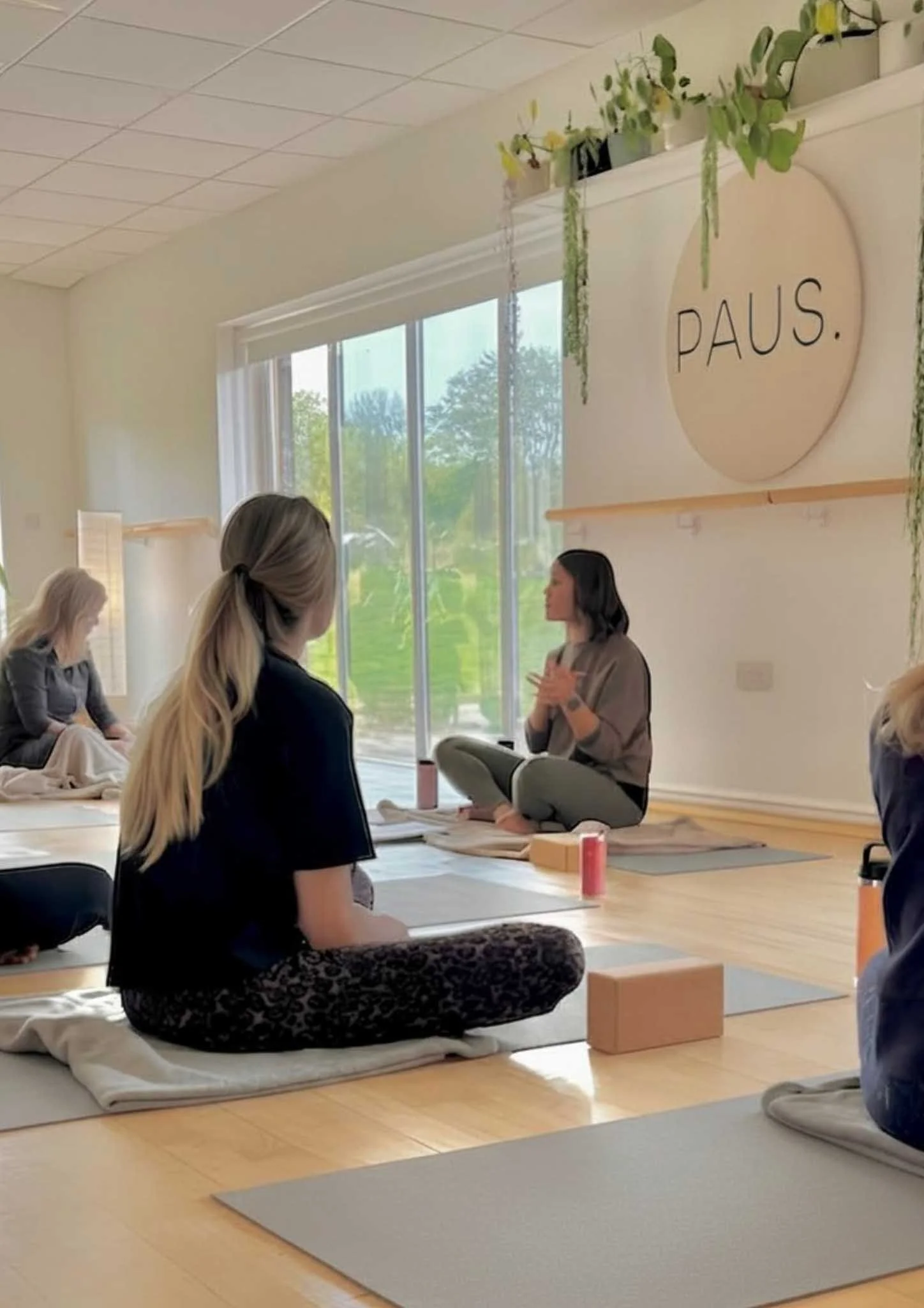 A woman leading a meditation or mindfulness class in a bright room with large windows. The participants are seated on yoga mats or cushions, facing her, with some in a cross-legged position. The room is decorated with plants and a large sign that say