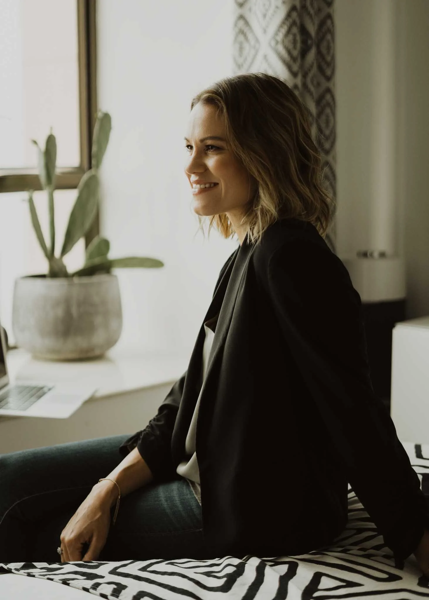 A woman with shoulder-length wavy hair sitting on a bed, dressed in a black blazer and jeans, smiling softly, with a window and a decorative wall hanging behind her, a plant on the windowsill, and a zebra-patterned blanket in the foreground.