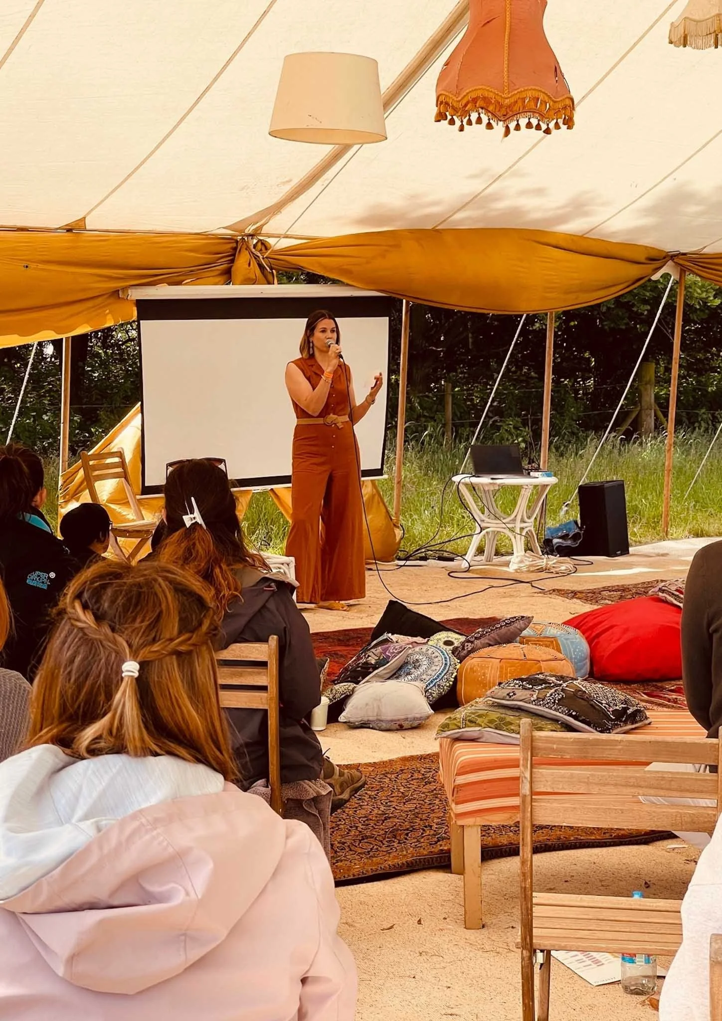 A woman in burnt orange jumpsuit stands and speaks into a microphone inside a large, open-sided tent with yellow drapes. The tent has decorative hanging lamps and lampshades on the ceiling. Audience members sit on the ground on pillows and cushions, some with braided hair, and face the speaker. There is a large white projection screen behind her, and outdoor greenery is visible beyond the tent.