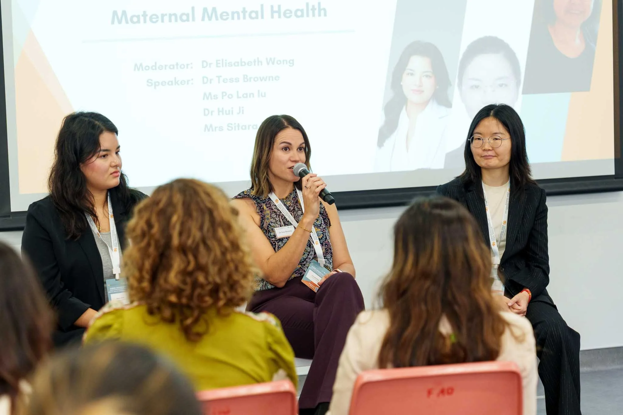 A panel discussion at a conference on Maternal Mental Health, with three women seated and speaking to an audience. A slide on the screen behind them lists the session details and speakers' names.