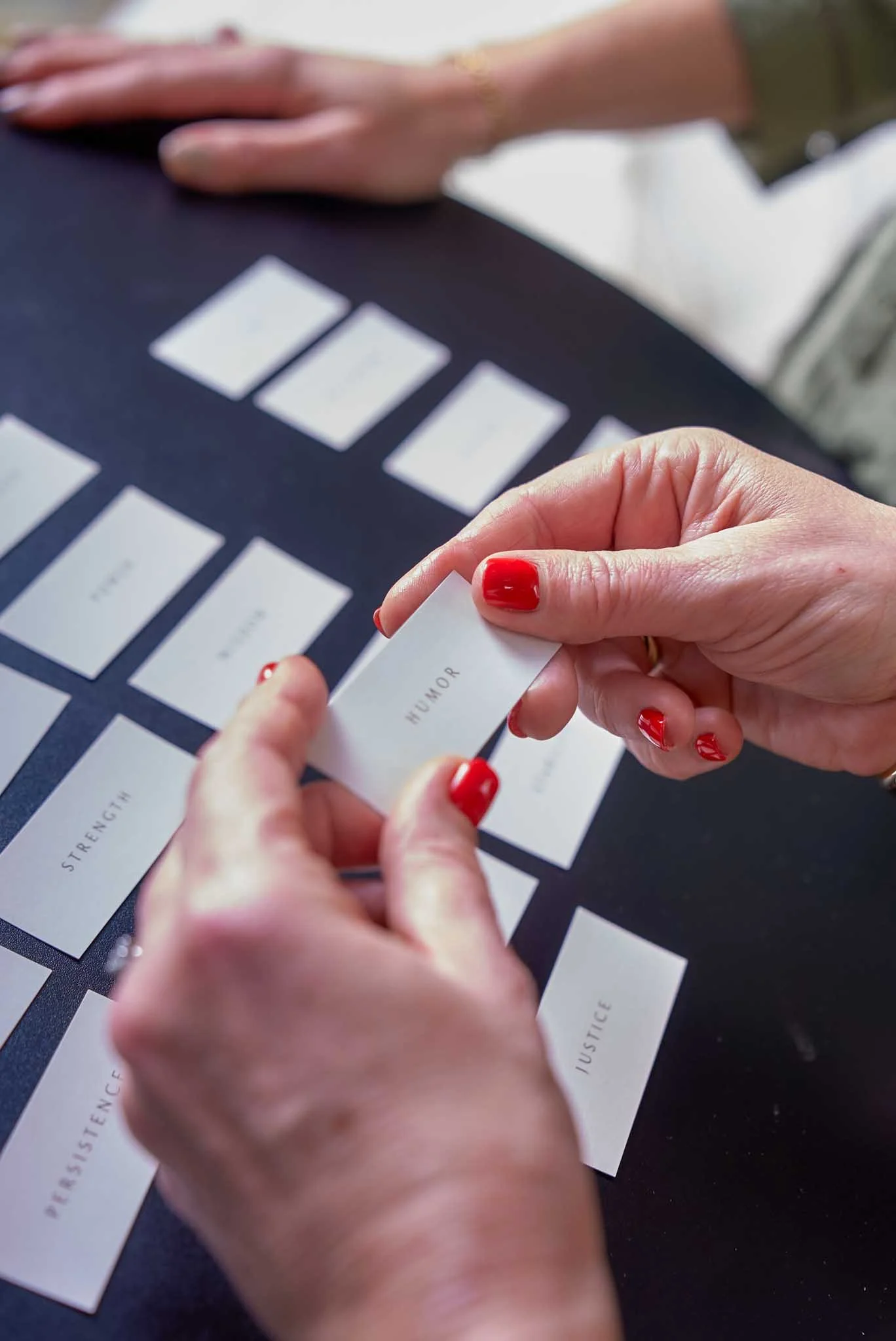 Close-up of a person's hands with red nail polish, holding a small card labeled 'HUMOR' over a grid of white cards with black text, on a dark surface.