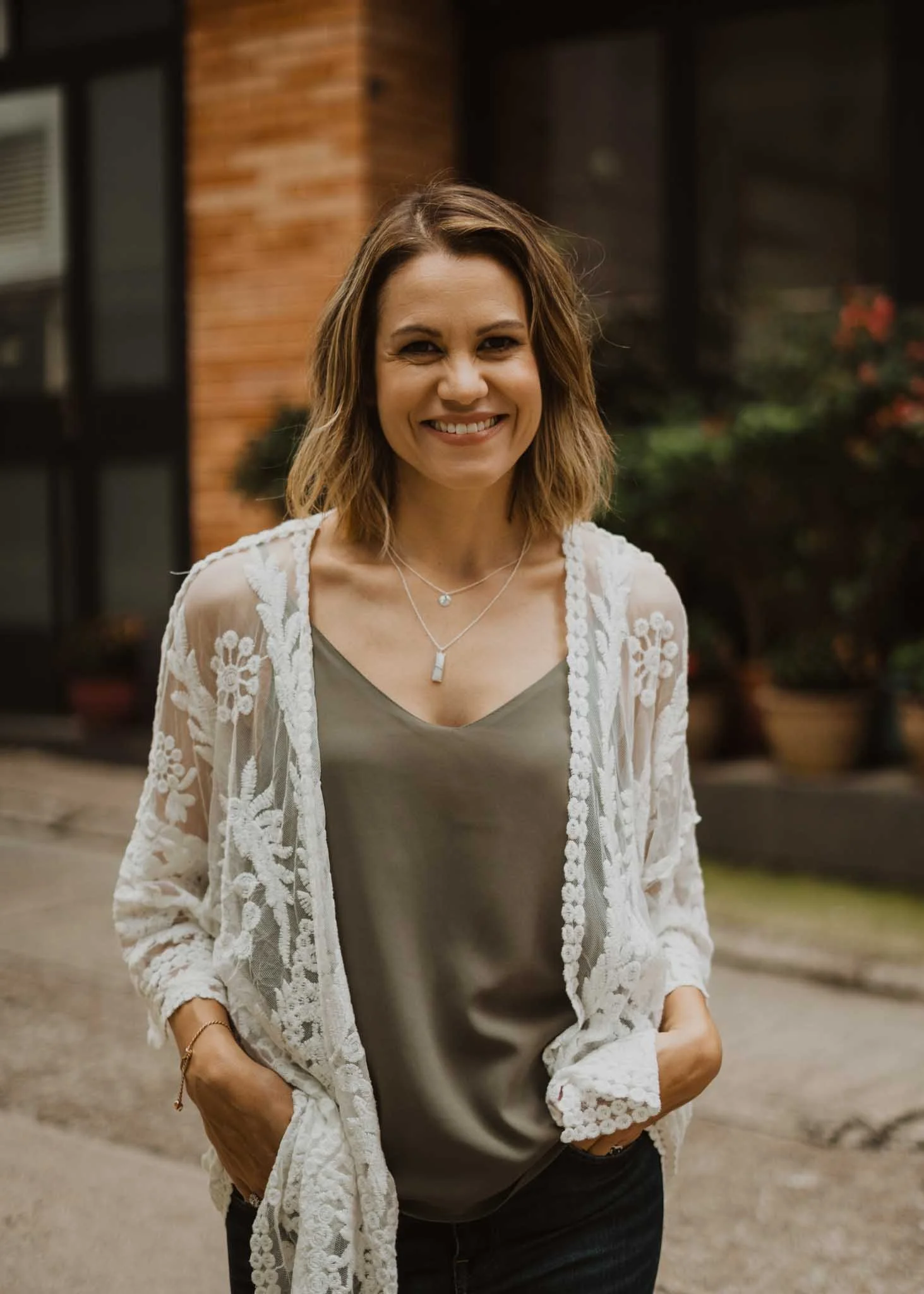 A woman with shoulder-length brown hair, smiling, standing outdoors in front of a brick building and greenery.