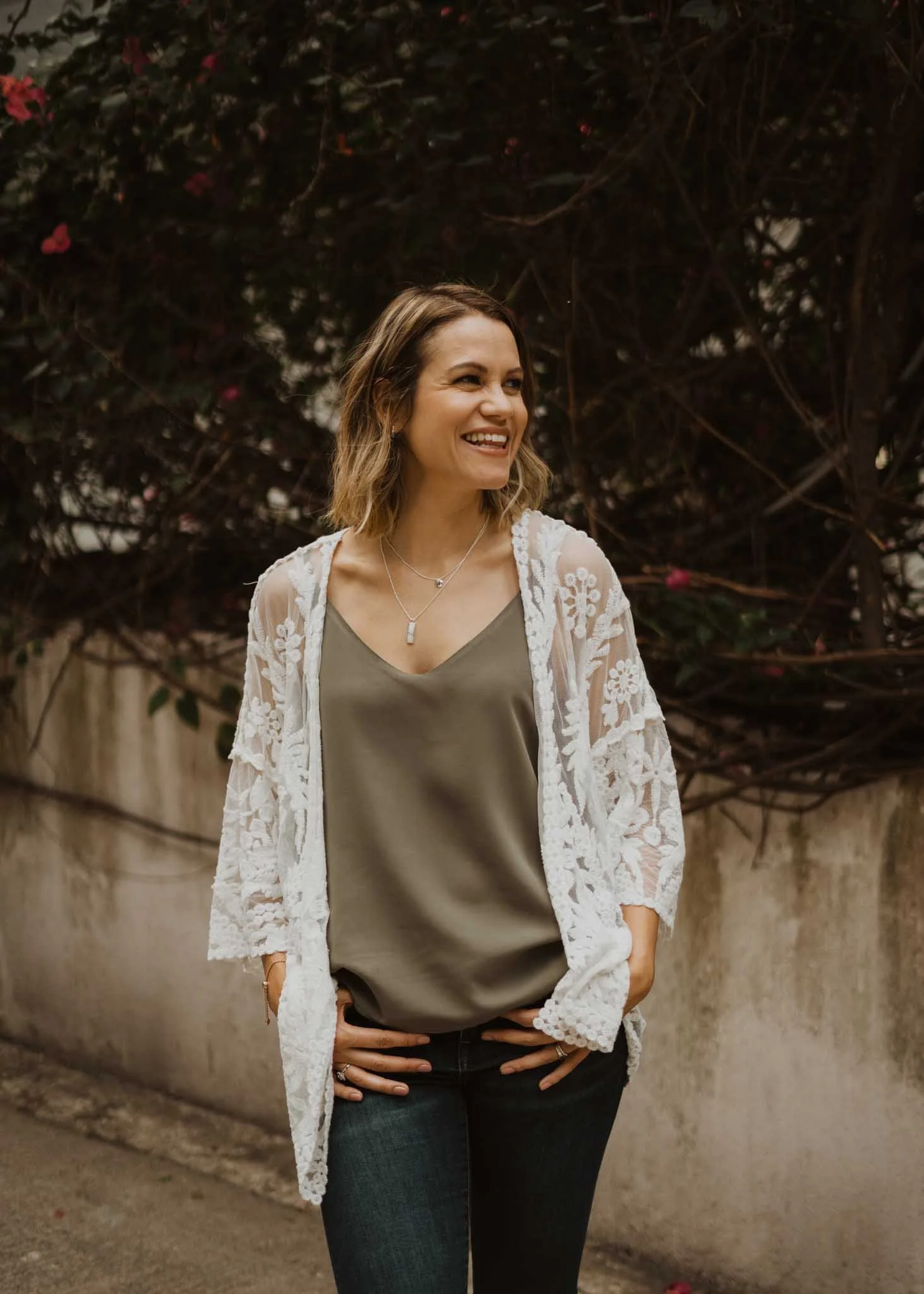 A woman with shoulder-length wavy hair smiling, wearing a green top, layered necklaces, and a white lace cardigan. She stands outdoors in front of a bush with dark leaves and pink flowers, with her hands on her hips.