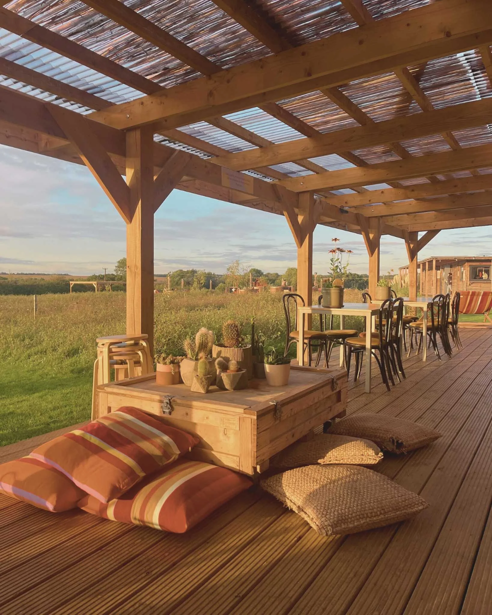 Outdoor patio with wooden pergola, seating table, chairs, striped cushions, and potted cacti, overlooking a rural landscape.