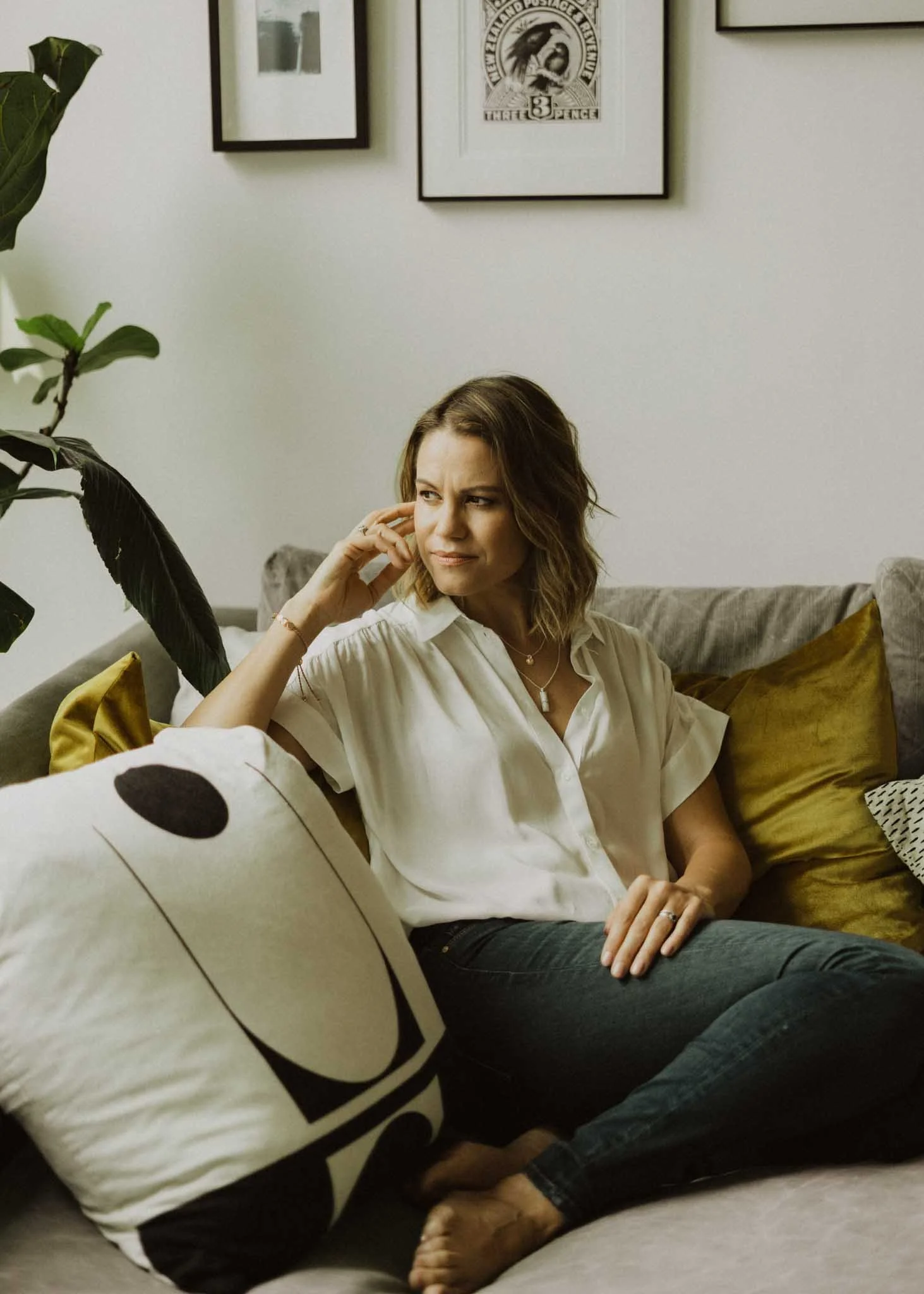 A woman with brown hair sitting on a sofa, looking to the side with a thoughtful expression. She is wearing a white blouse and dark jeans. The sofa has decorative pillows in yellow and brown tones. Behind her, framed artwork is hanging on a light-colored wall, and part of a large leafy plant is visible on the left side of the image.
