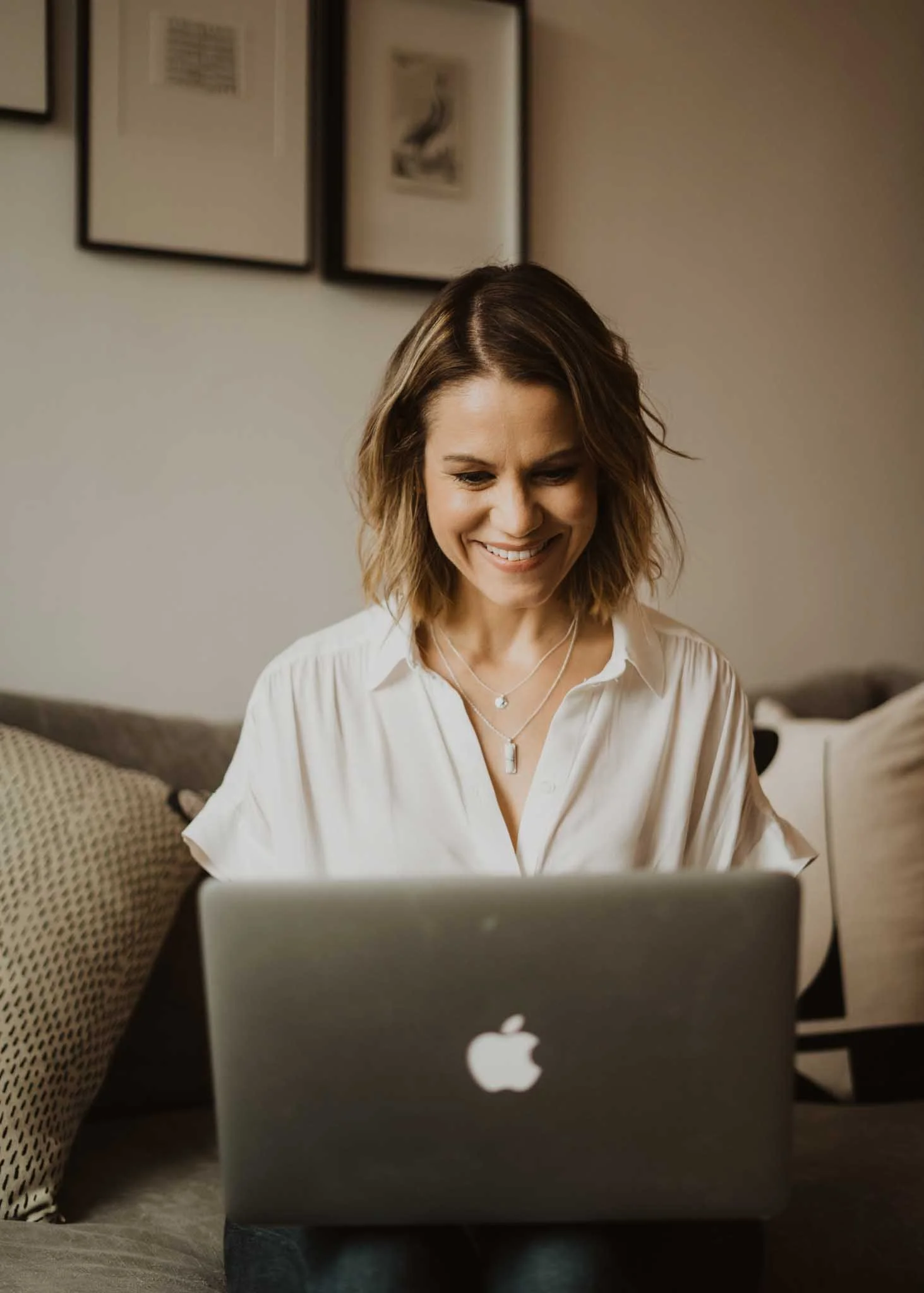 A woman with shoulder-length brown hair, smiling and wearing a white shirt, sitting on a beige sofa, working on a silver MacBook laptop with an Apple logo, in a room with framed artwork on the wall.