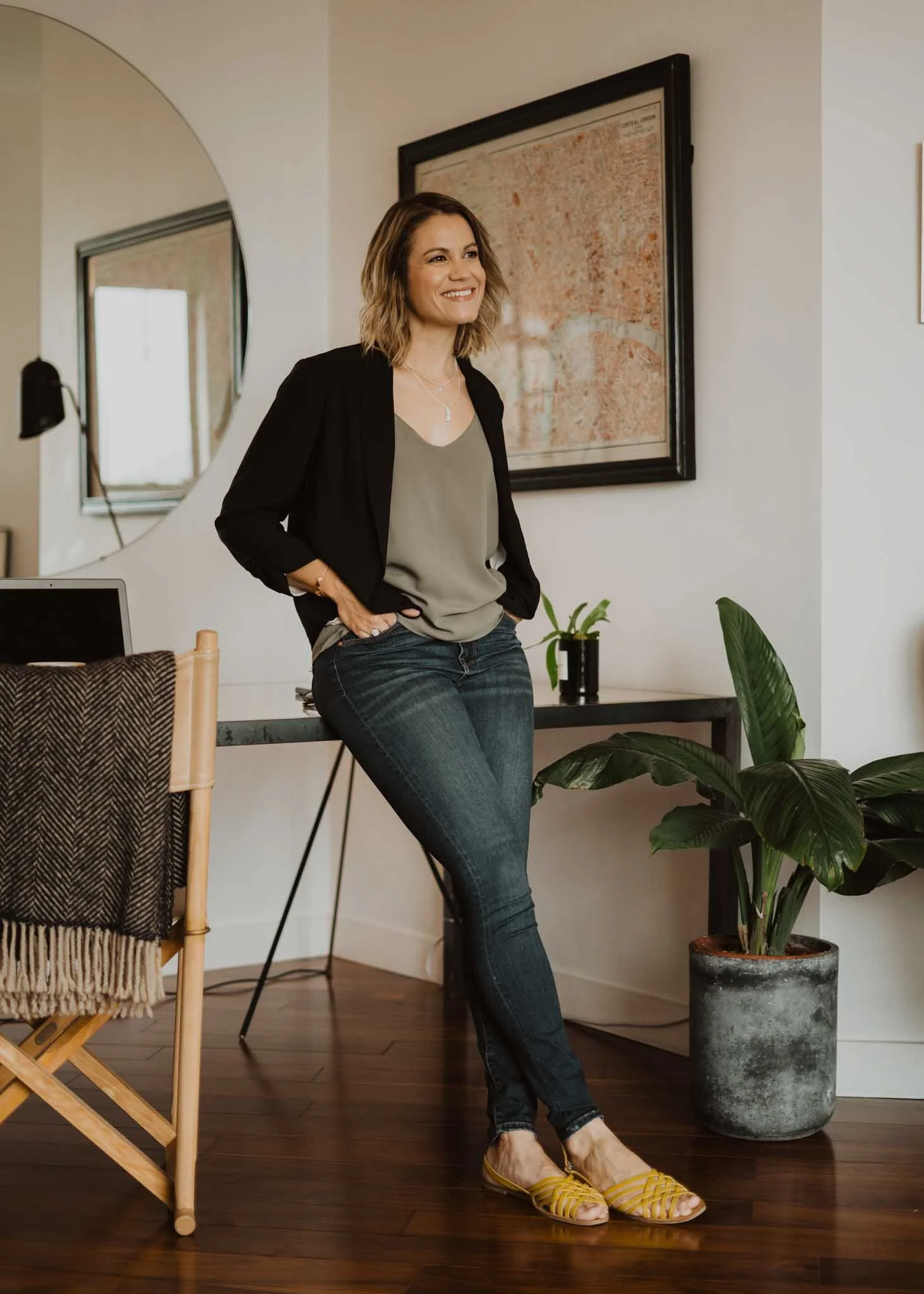 A woman with short, wavy hair, wearing a black blazer, gray top, and dark jeans, standing in a room with wooden floors, near a large potted plant and a framed map on the wall. She is smiling and wearing yellow shoes.