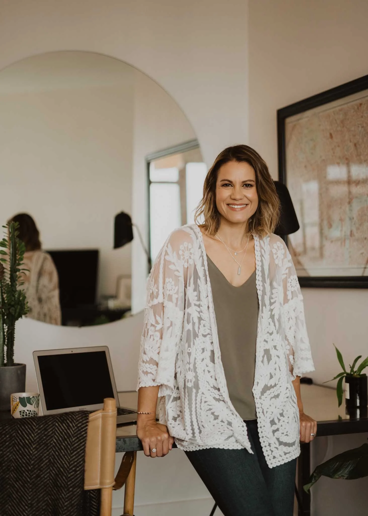 Woman standing in a modern office wearing a gray top and a white lace cardigan, smiling at the camera, with a desk, laptop, and plants in the background.