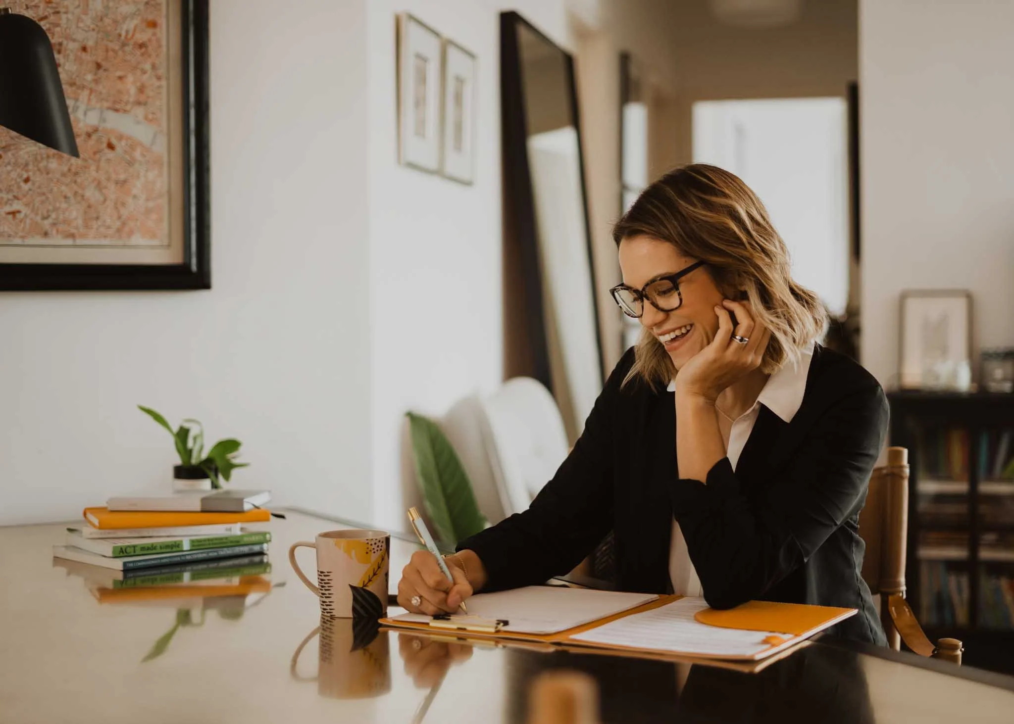 A woman with glasses and wavy hair sitting at a desk, smiling, writing on a pad, with a mug and a stack of books nearby. The background shows a wall with framed pictures and a window.