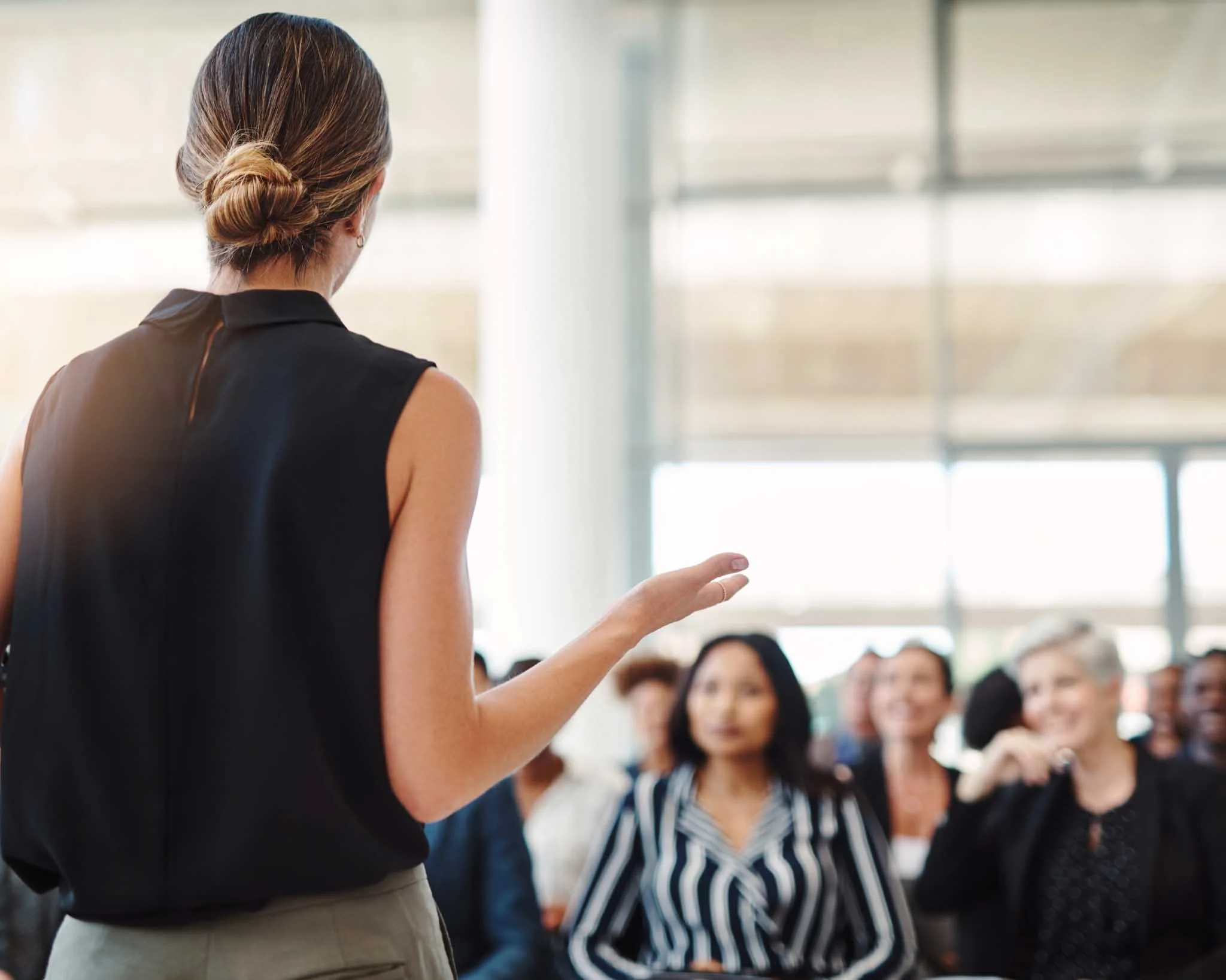 A woman speaking to a group of diverse women in an indoor setting with large windows, during a presentation or workshop.