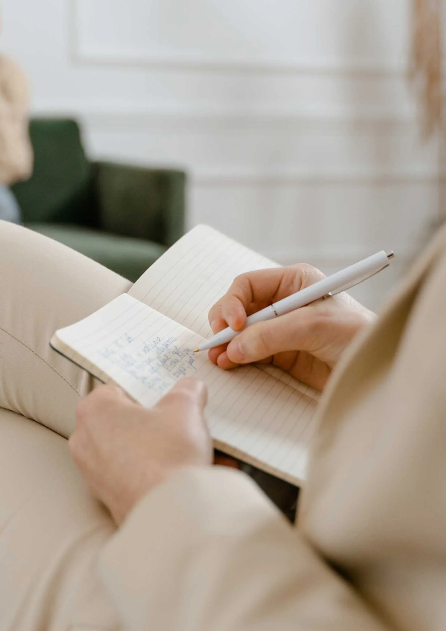 Close-up of a person writing in a lined notebook with a white pen, seated on a beige chair in a bright room.