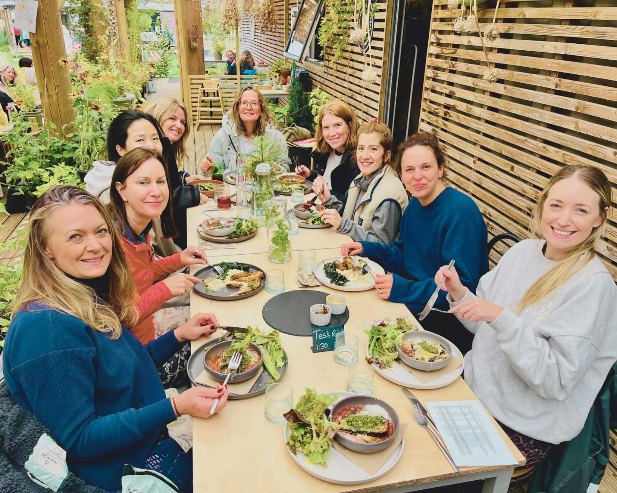 A group of women sitting around a table outdoors at a restaurant, enjoying a meal with plates of salads and pasta, surrounded by wooden fencing and green plants.